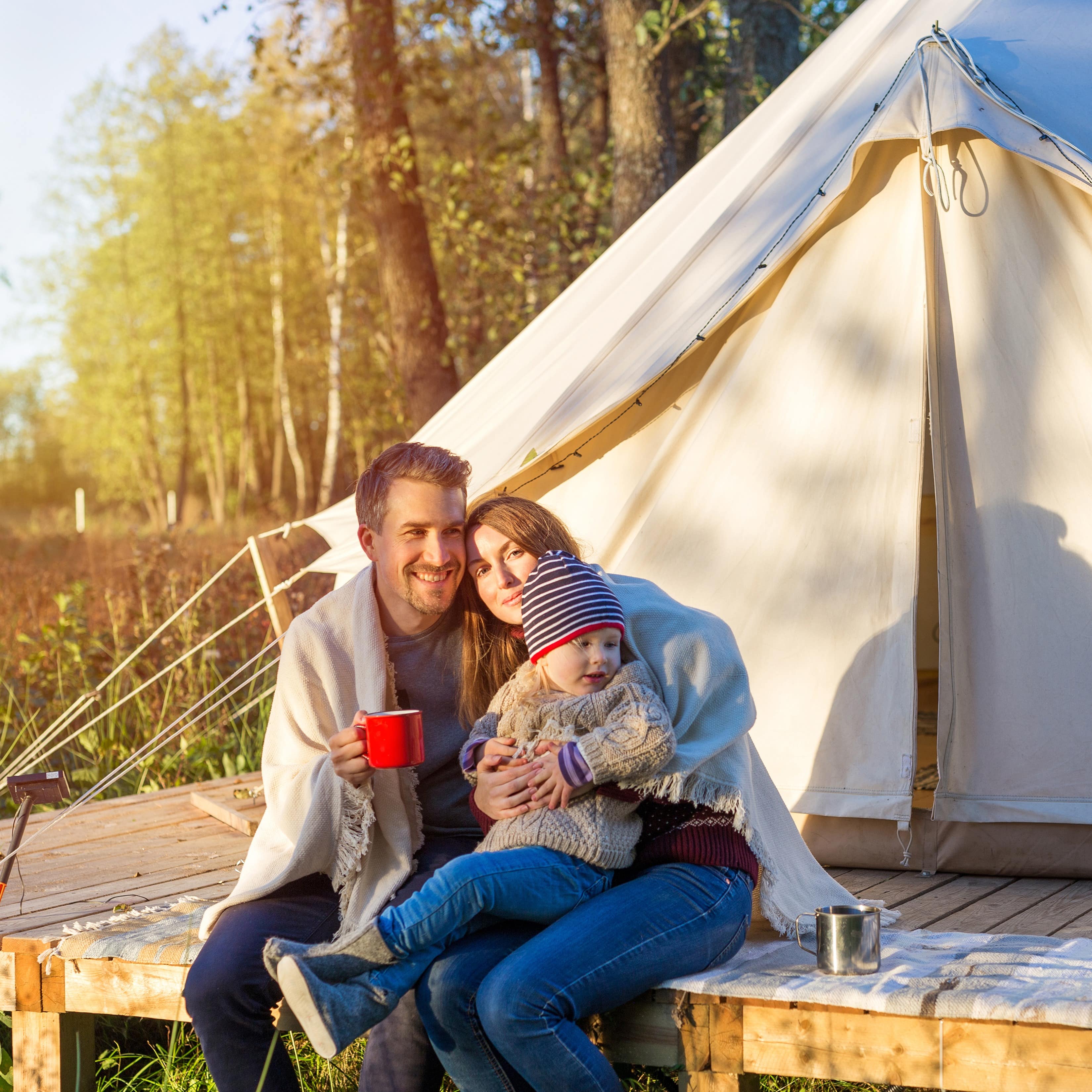 Vater, Mutter und kleines Kind sitzen in Decken eingehüllt auf einem Holzdeck vor einem Zelt. 