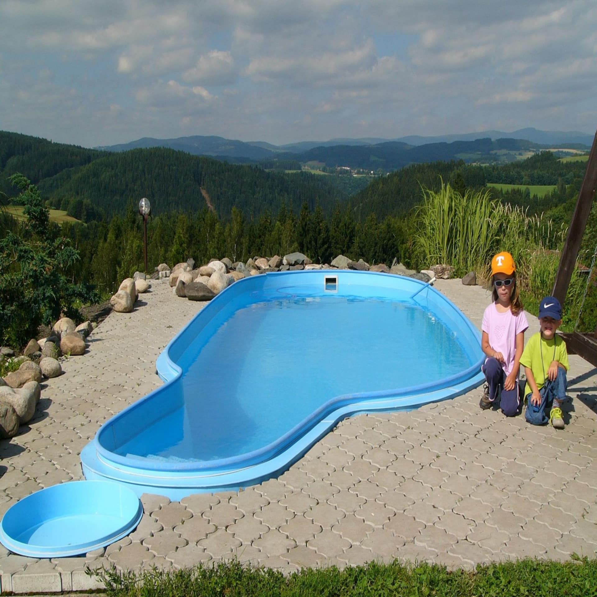 2 kleine Kinder neben einem Außenpool in einem Garten mit Panoramablick auf das grüne Umland.