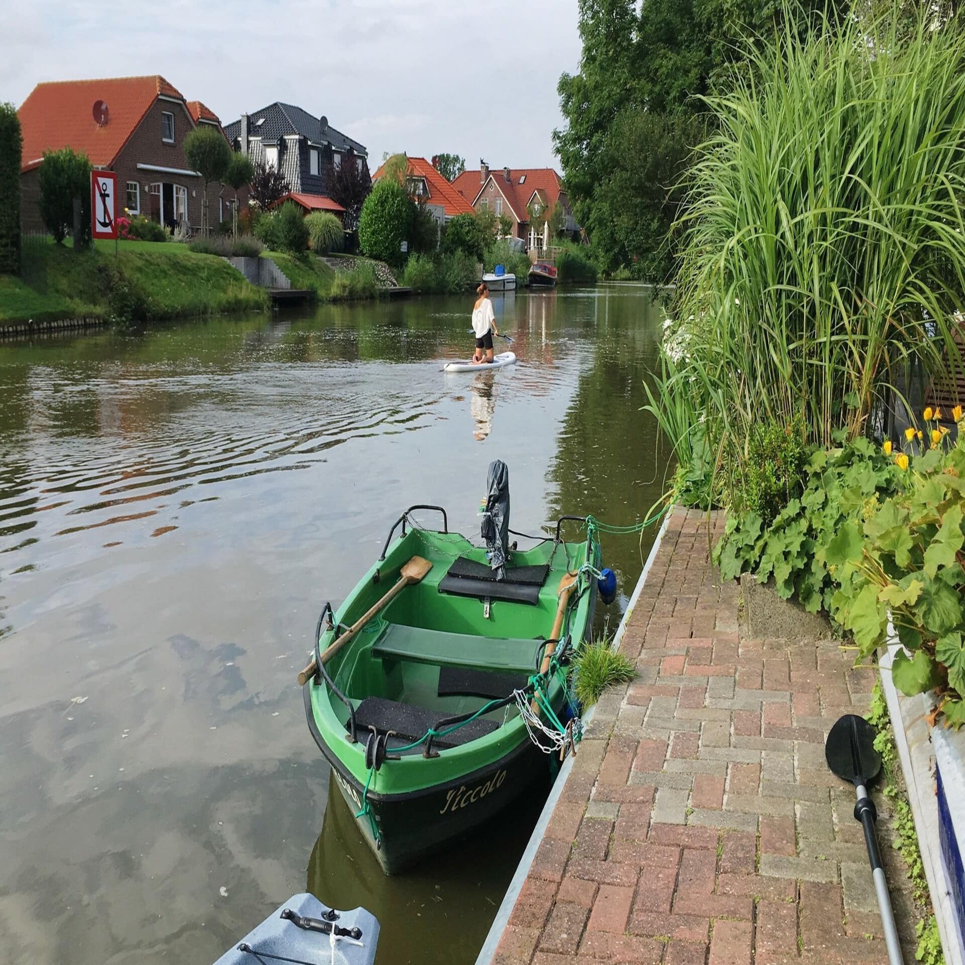 Grünes Ruderboot an einem Steg, eine Frau auf einem SUP und Häuser direkt am Wasser. 