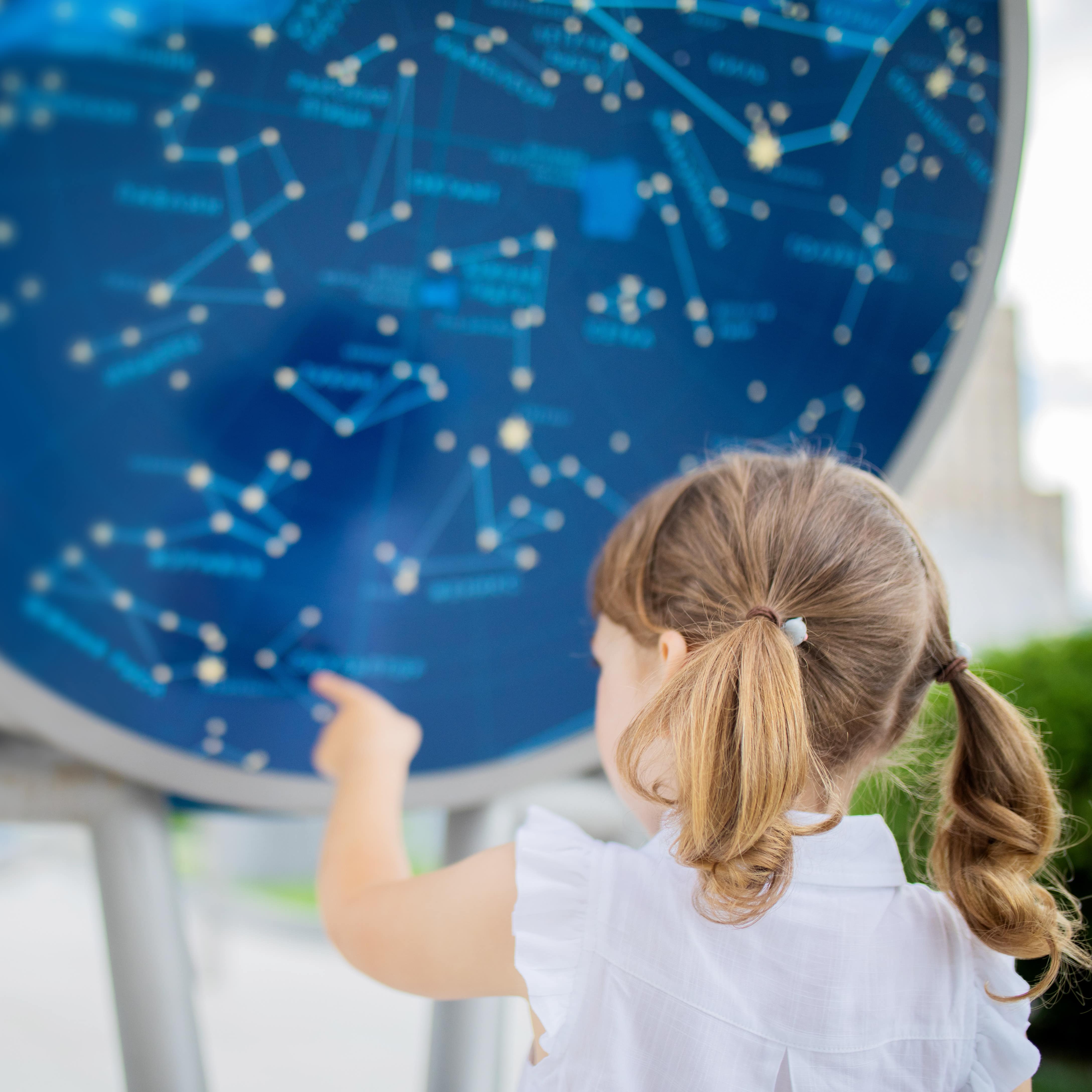 Rückenansicht: Ein kleines Mädchen mit Zöpfen zeigt auf Sternbilder in einer Ausstellung im Planetarium.