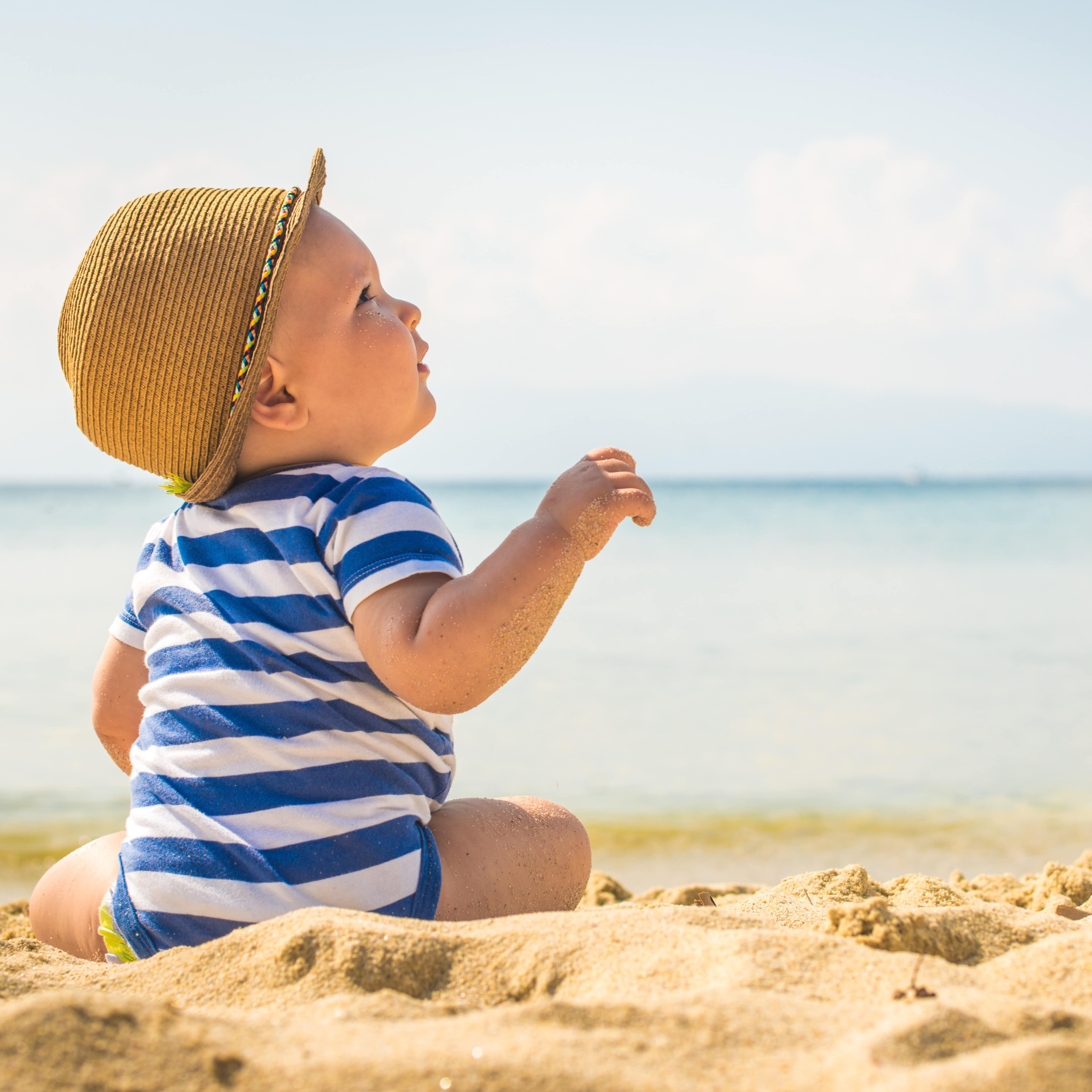  Ein Baby im gestreiften Strampler und Strohhut sitzt am Strand und schaut nach rechts oben.