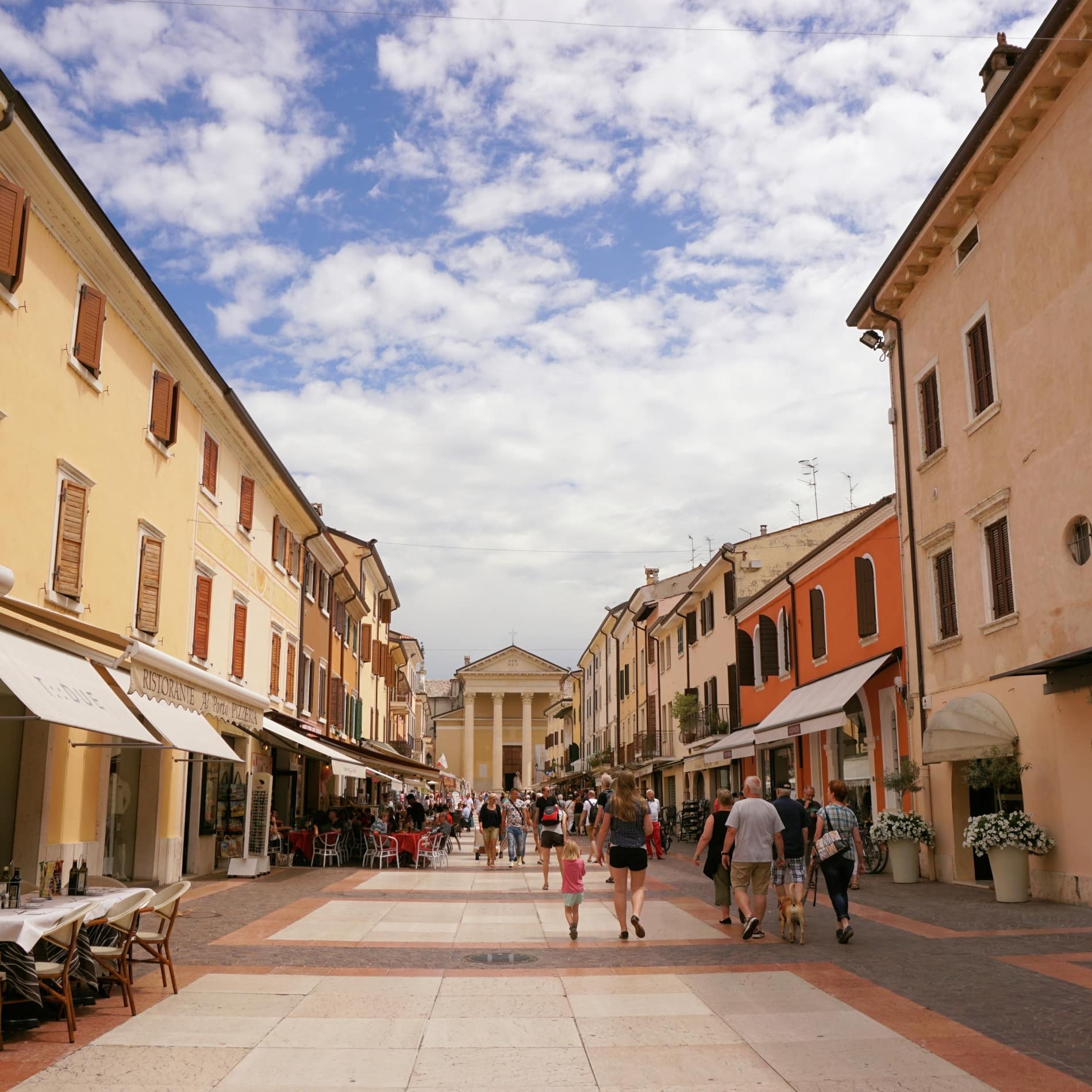 Touristen schlendern durch eine Fußgängerzone in Bardolino, im Hintergrund die Kirche San Nicolò mit Kolonnade