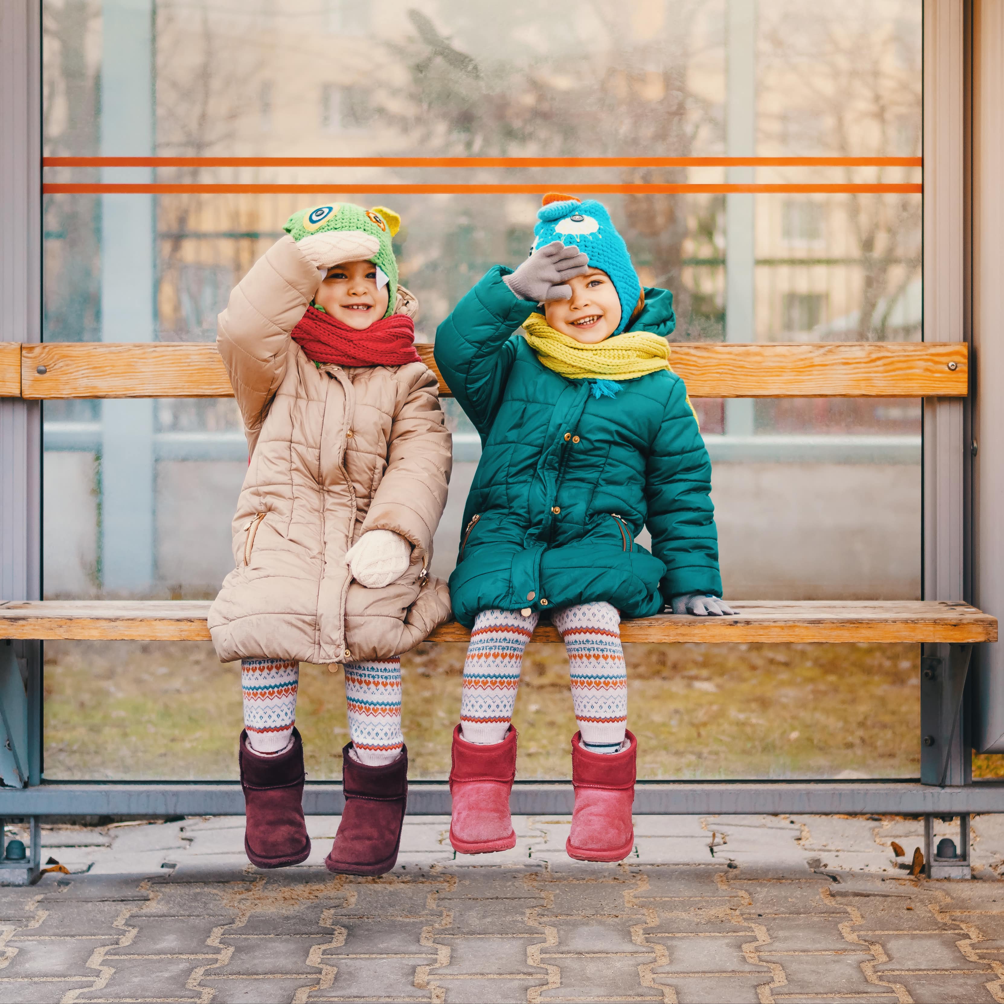 2 Mädchen in Winterkleidung sitzen auf einer Bank an einer Bushaltestelle. 