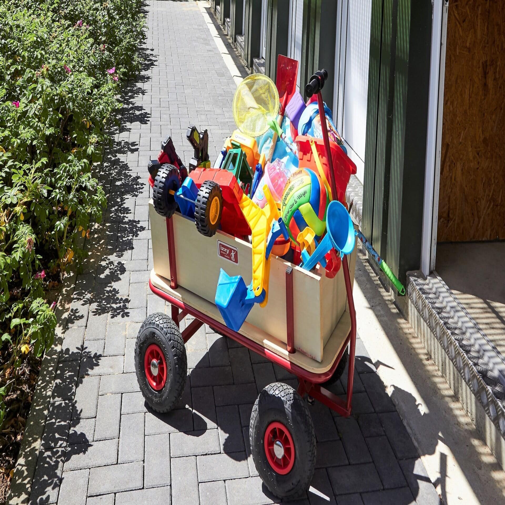 Ein Bollerwagen voller Strandspielzeug auf einem gepflasterten Weg neben einem Haus. Die Sonne scheint.