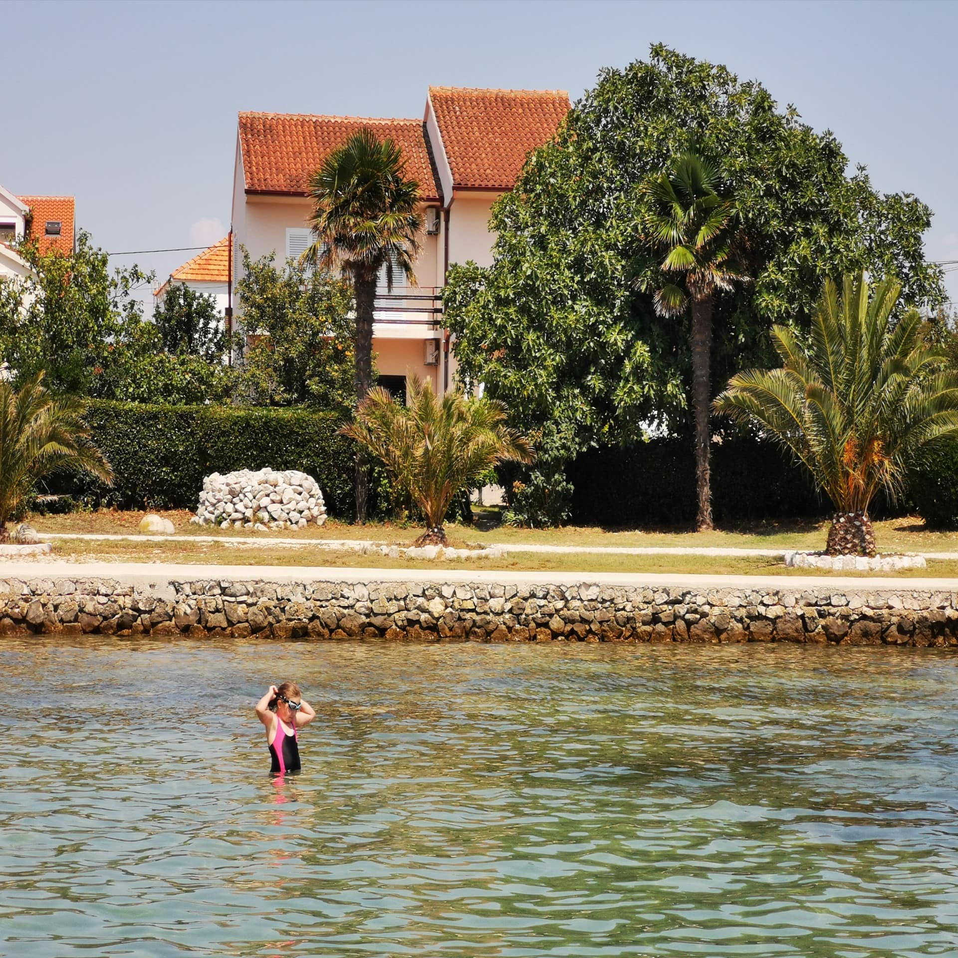 Mädchen im Wasser an einer Badestelle in Kroatien. Um Ufer Häuser. Die Sonne scheint.