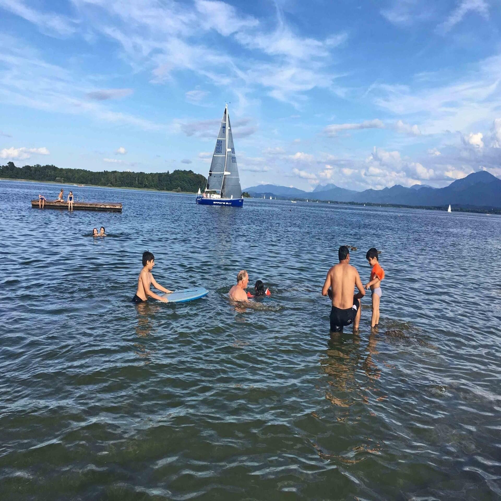Erwachsene und Kinder schwimmen im Chiemsee, ein Segelboot ist zu sehen.