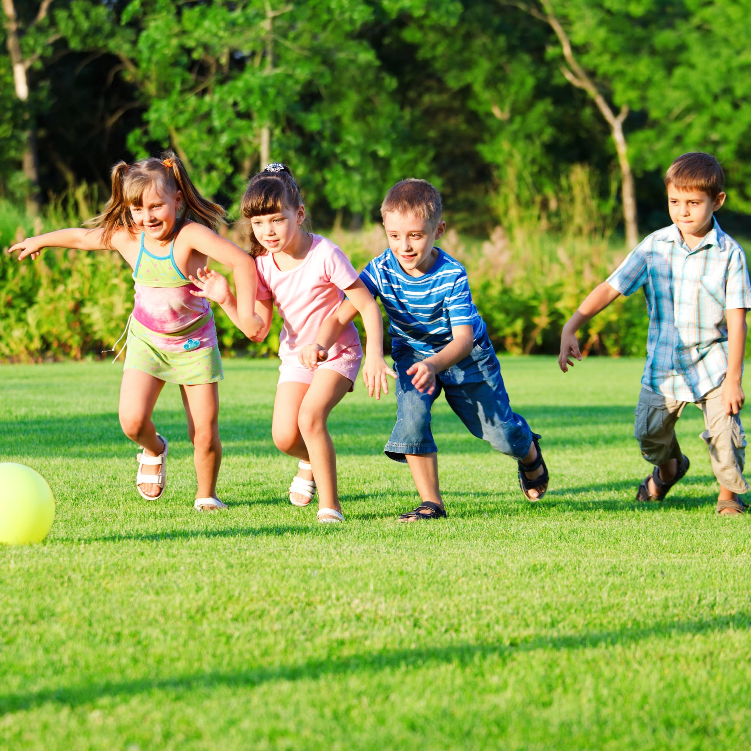 2 Jungs und 2 Mädchen in Sommerkleidung spielen mit einem Ball auf einer Wiese.