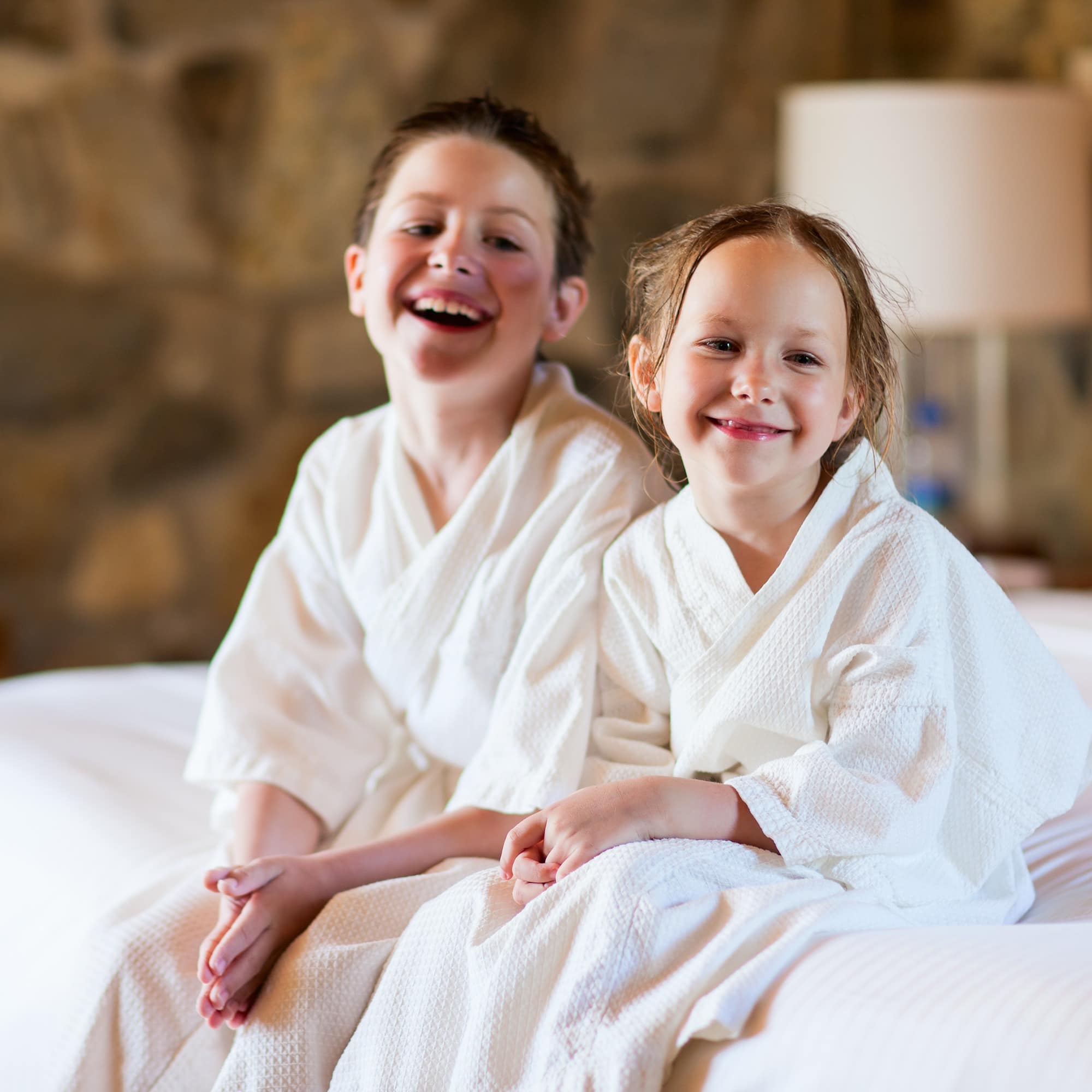 Bruder und Schwester in weißen Bademänteln sitzen auf einem weißen Hotelbett.