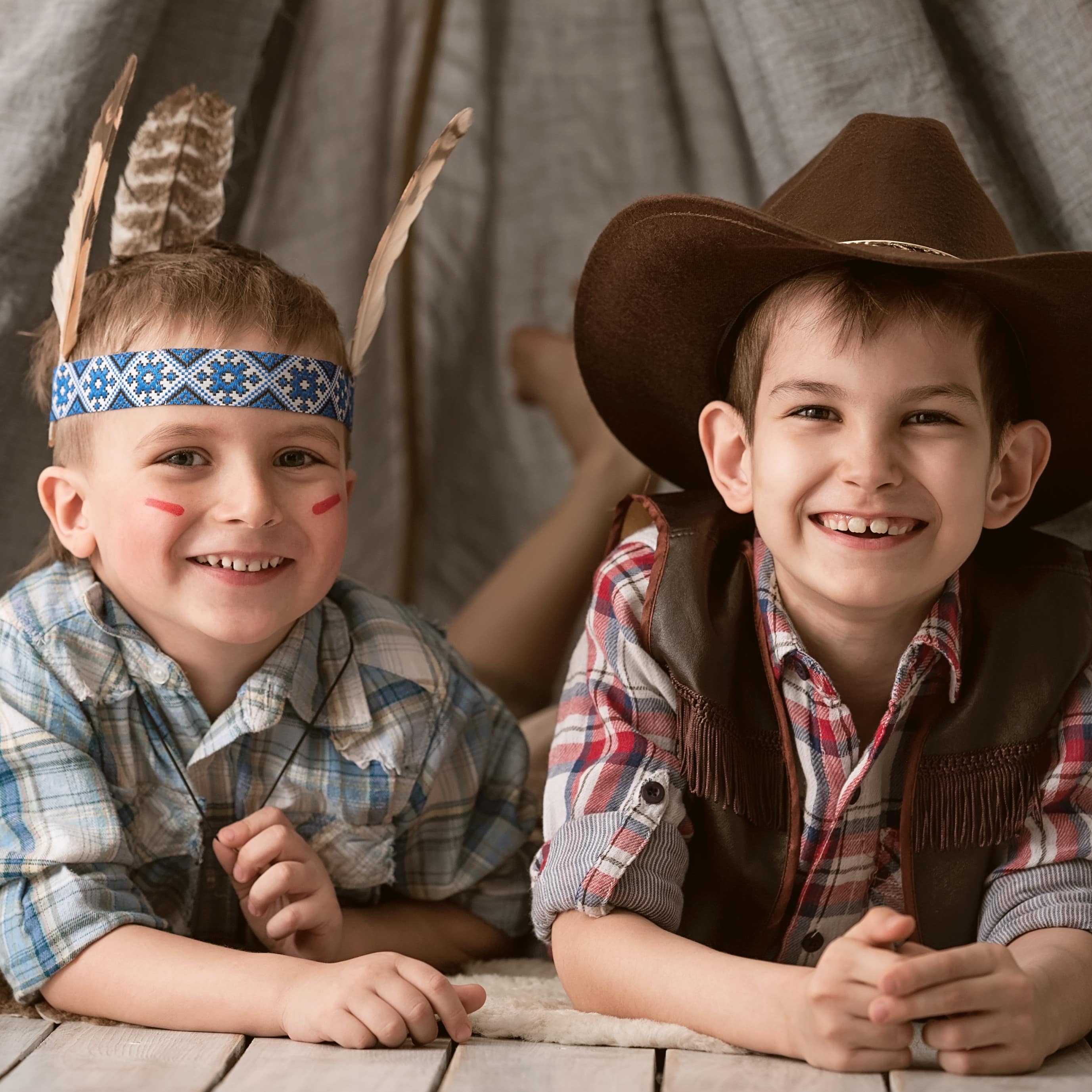 2 Jungen in Cowboy und Indianer-Verkleidung  liegen vor einem Zelt und lachen in die Kamera.