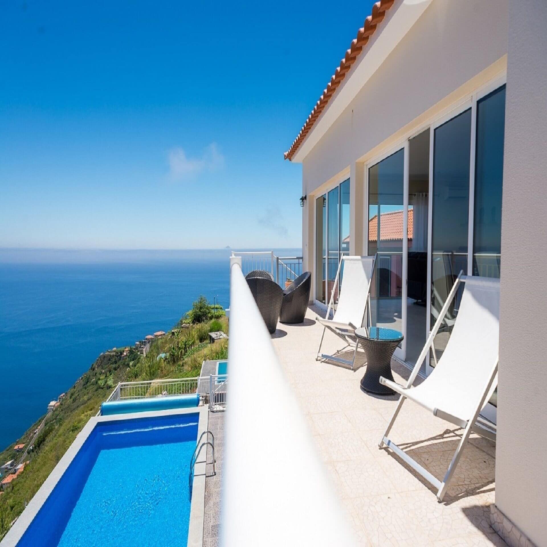 Balkon mit Meerblick eines weißen Hauses, darunter ein Pool auf einem Hügel. 