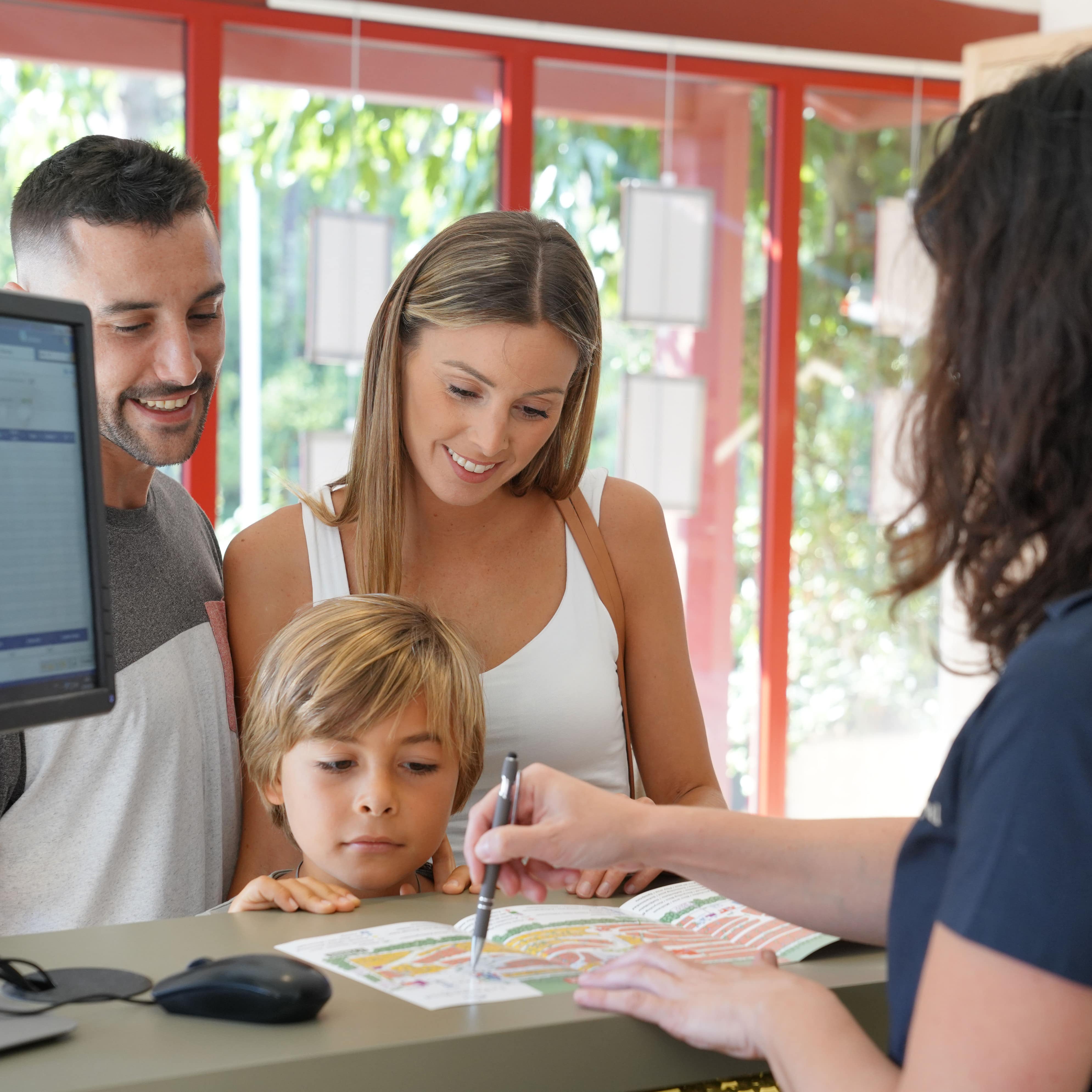 Eine Familie mit Sohn an der Rezeption eines Hotels, die Rezeptionistin zeigt etwas auf einem Stadt-plan.