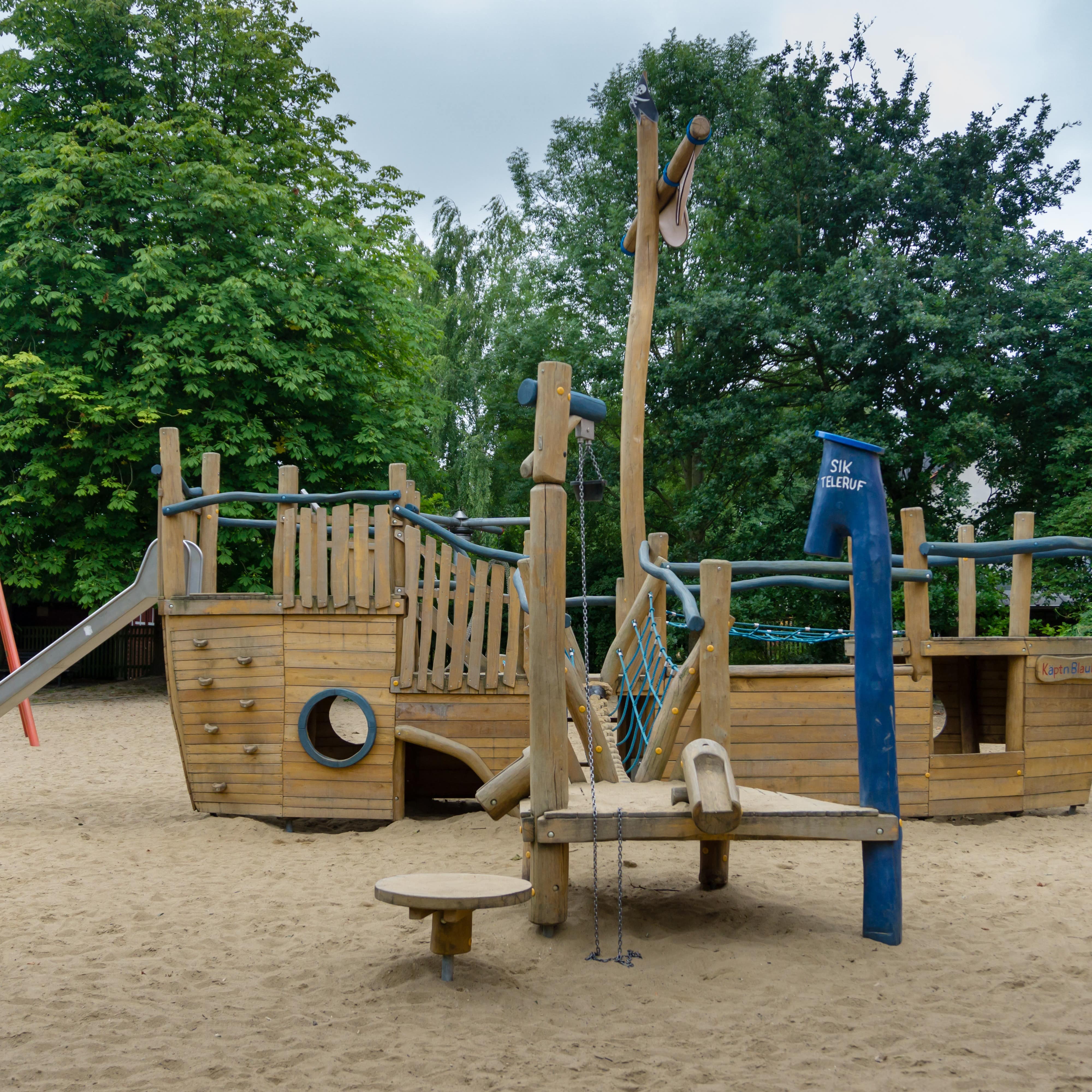 Sandspielplatz mit einem Holzschiff und Klettergerüst, dahinter Bäume.