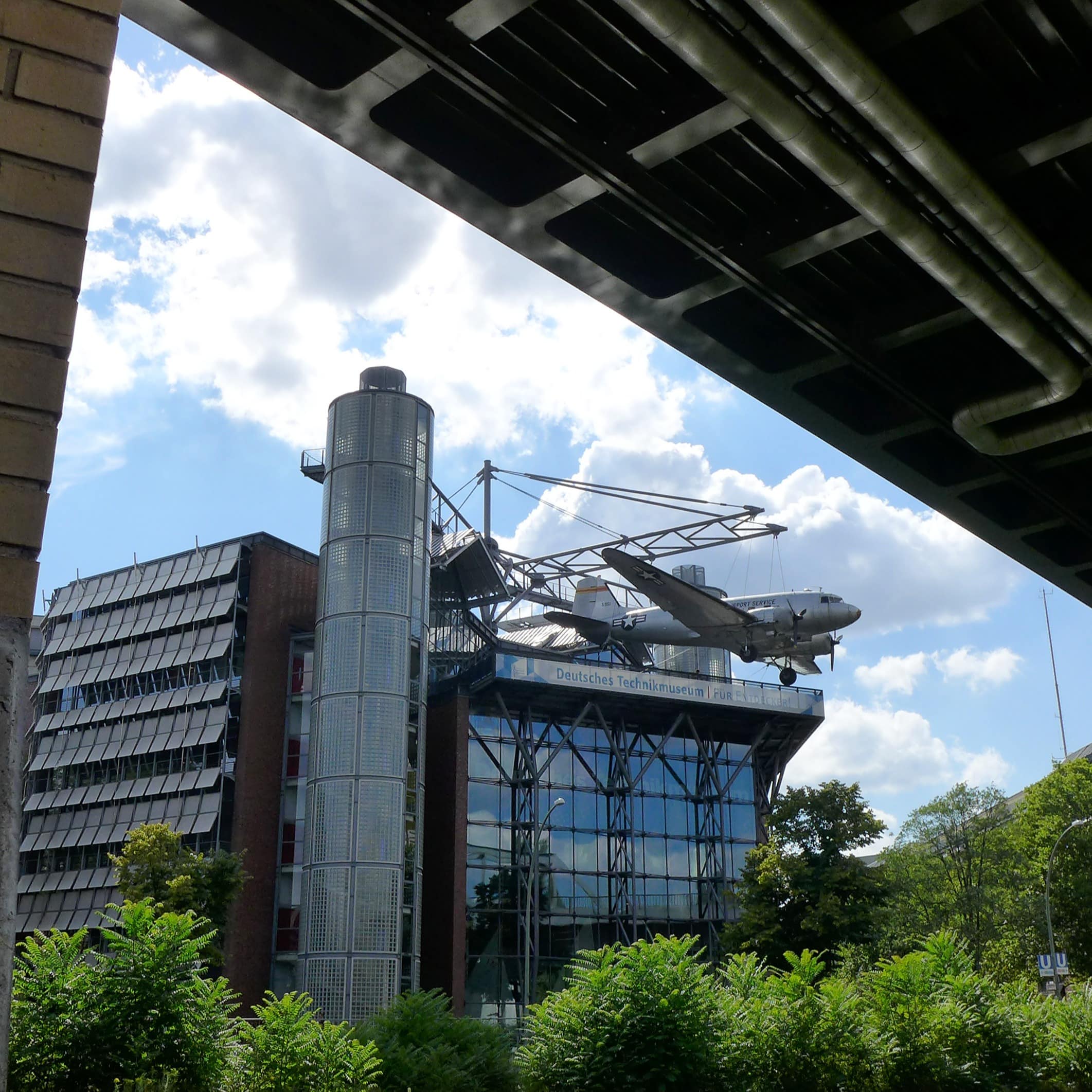 Blick unter einer Brücke hindurch auf das Gebäude des Technikmuseums über dem ein Flugzeug hängt. 