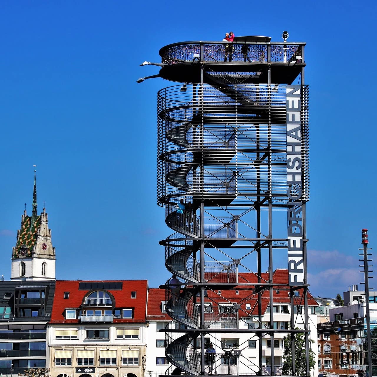 Blick auf eine hohe Aussichtsplattform, die über eine Treppe erreichbar ist, Häuser von Friedrichshafen im Hintergrund. 