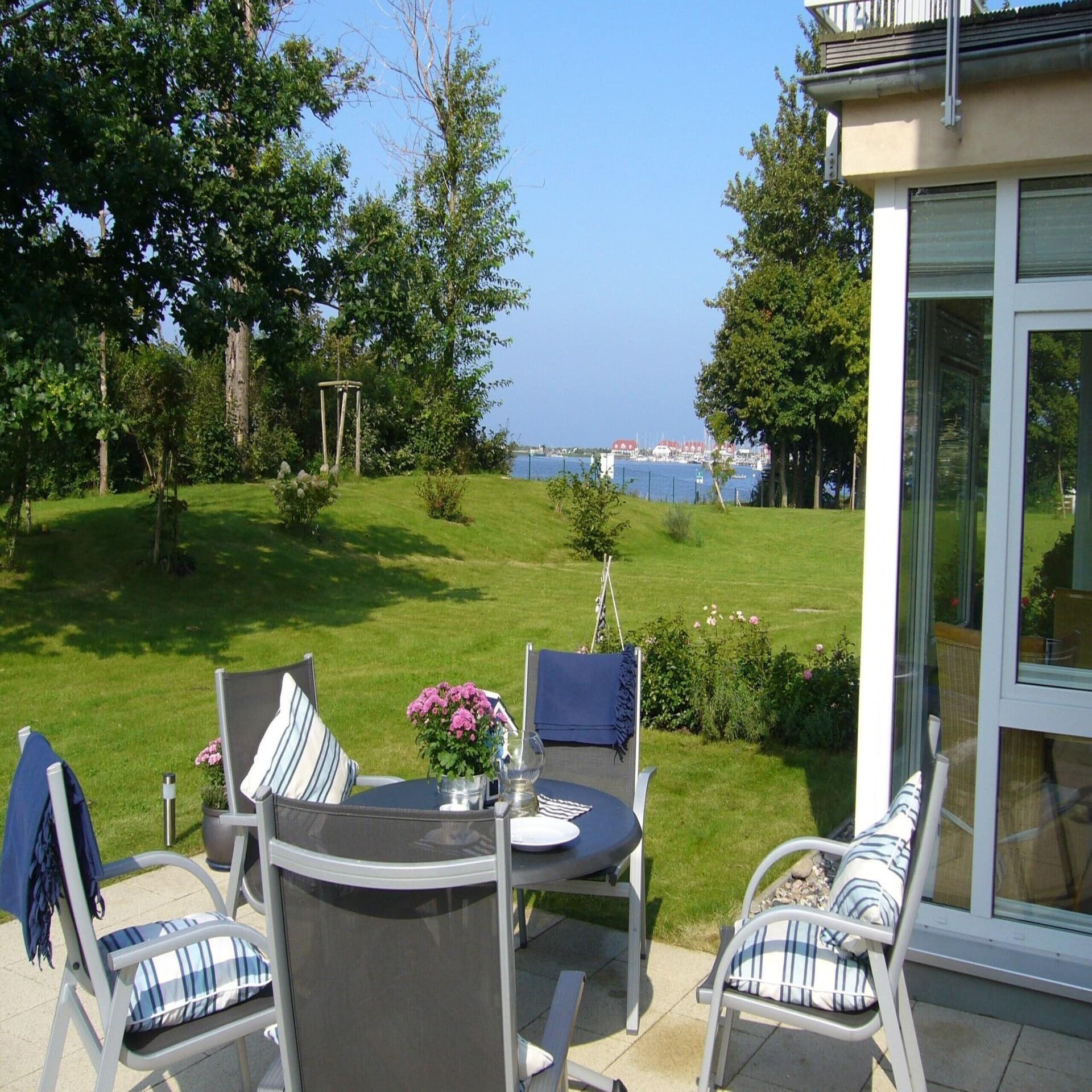 Terrasse mit Tisch und 5 Stühlen in einem sommerlichen Garten mit Blick auf das Wasser und Häuser in der Ferne.