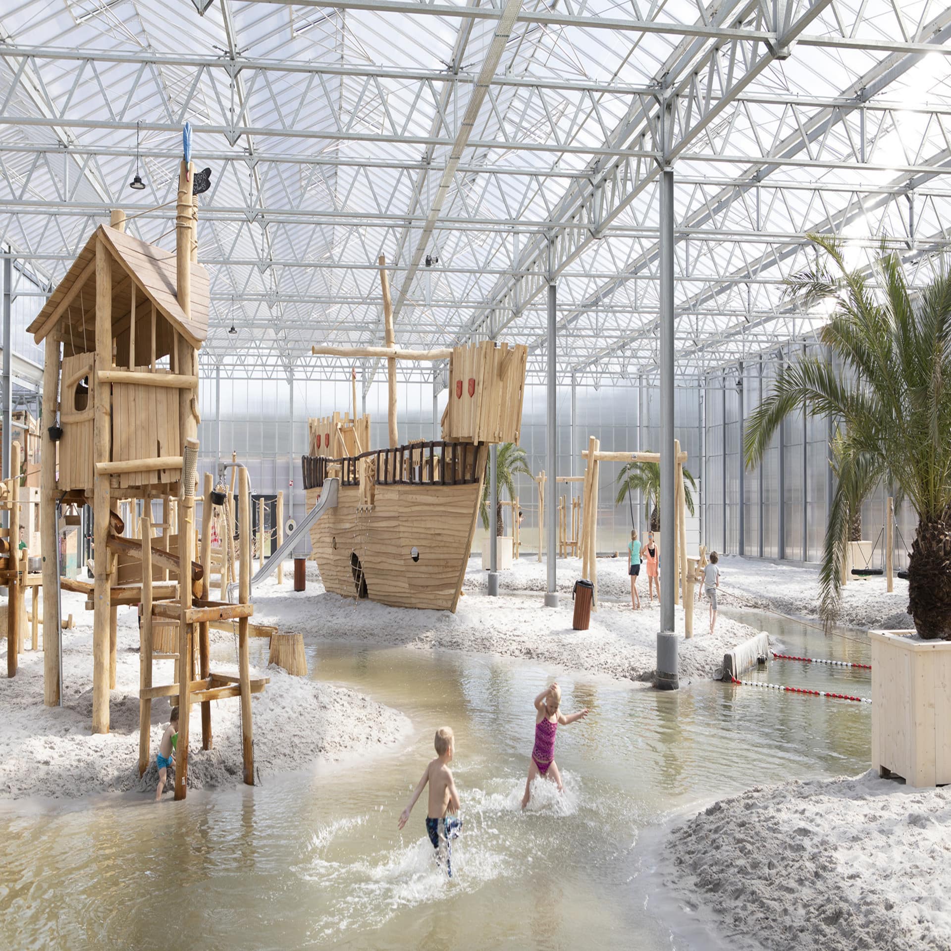 Kinder spielen in einer überdachten Sand- und Wasserlandschaft mit Abenteuerspielplatz.