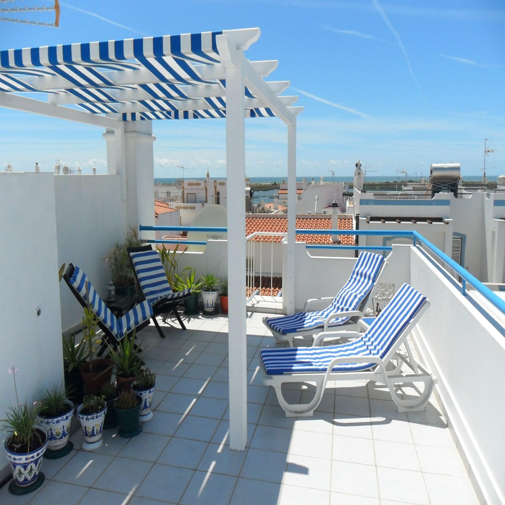 Dachterrasse mit Sonnenliegen und Sonnenschutz sowie Blick über Häuserdächer auf das Meer. 