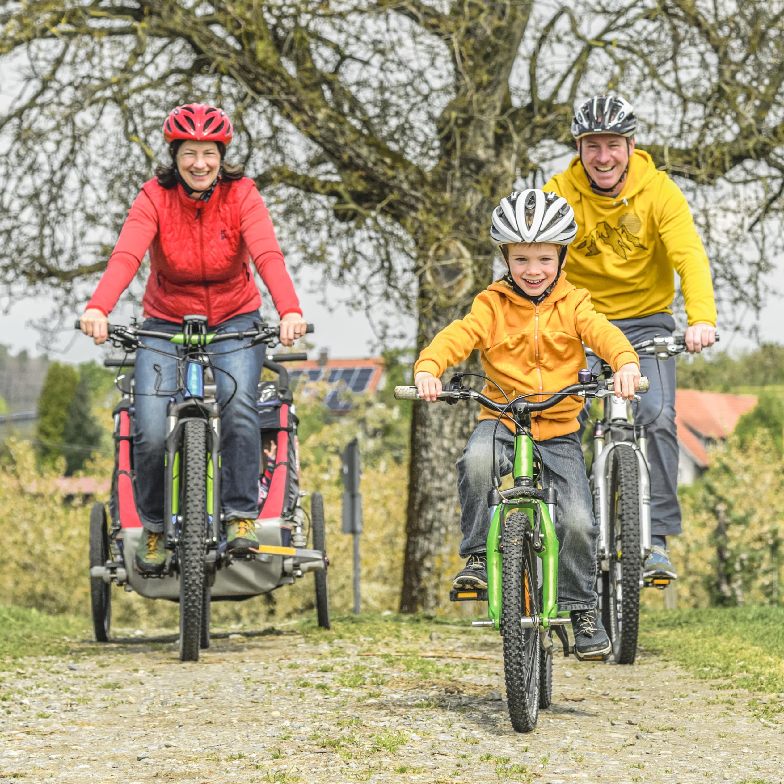 4-Köpfige Familie auf Fahrradtour im Frühling. Der Junge fährt vorne, die Mutter zieht einen Anhänger mit Baby drin.