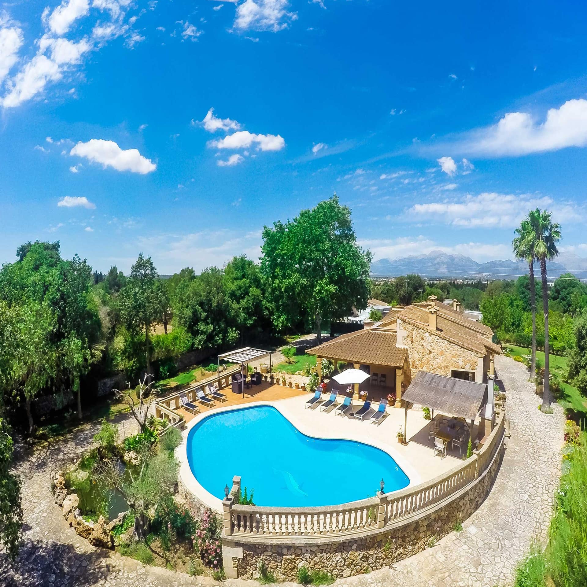 Blick von oben auf eine kleine Finca mit Pool mitten im Grünen. Die Sonne scheint.