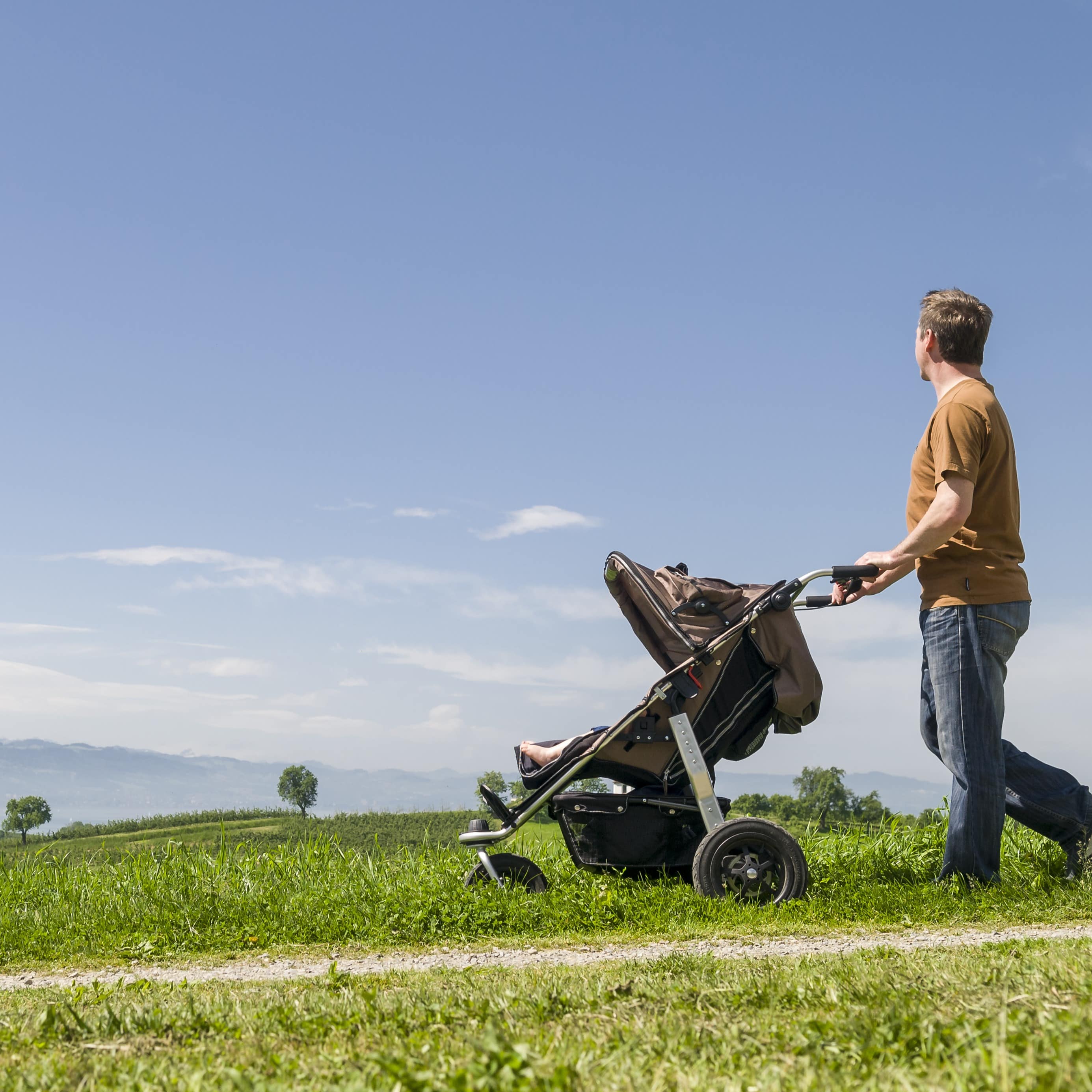 Ein junger Vater schiebt einen Kinderwagen durch einen ländlichen Pfad entlang, im Hintergrund Häuser.