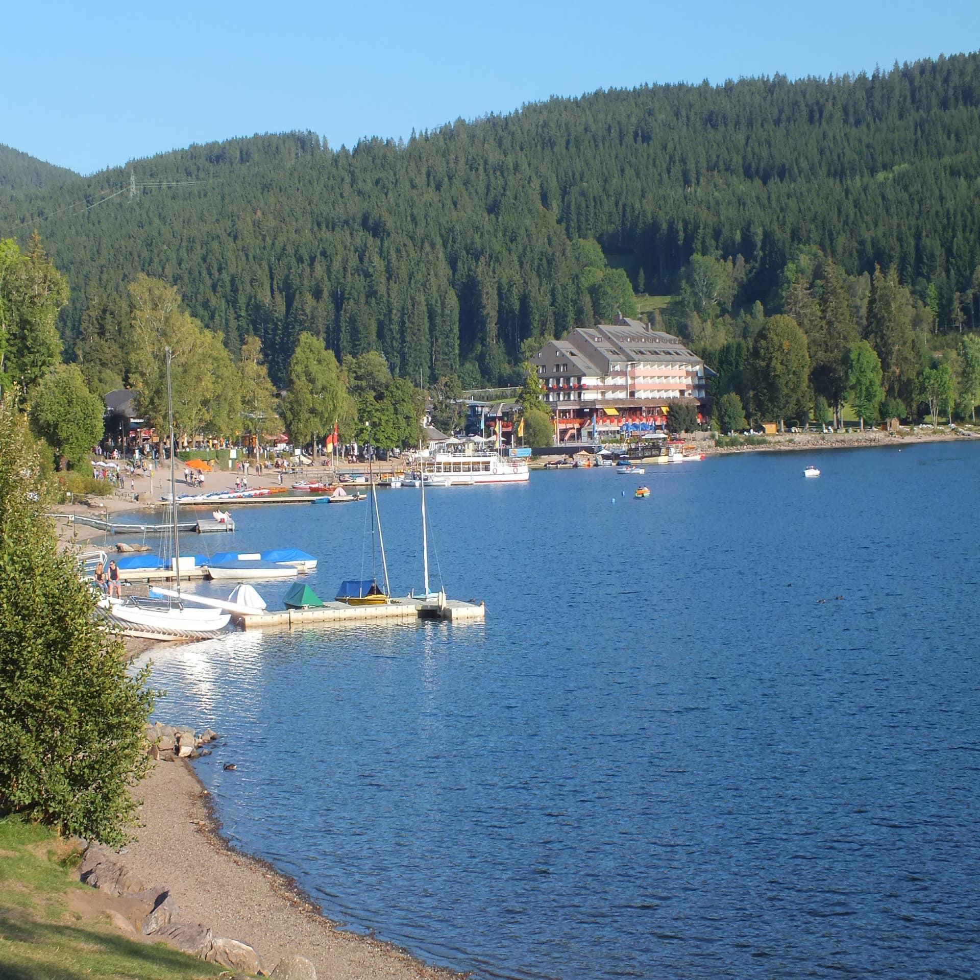 Blick über den Titisee und das Ufer an dem einiger Boote liegen. Im Hintergrund ein größeres Gebäude.  