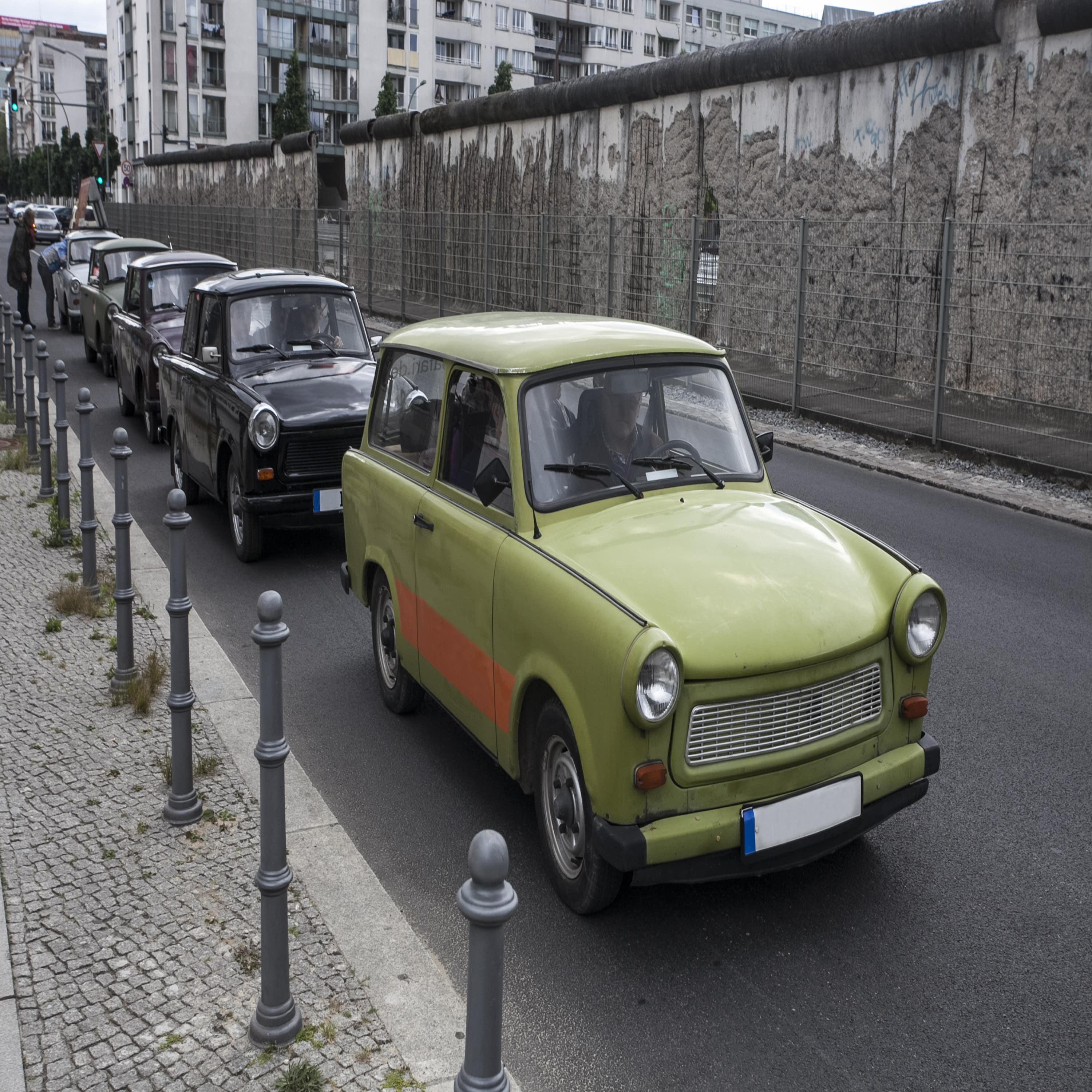 Ein grau-grüner Trabbi auf einer Straße, dahinter schwarze Trabbis.