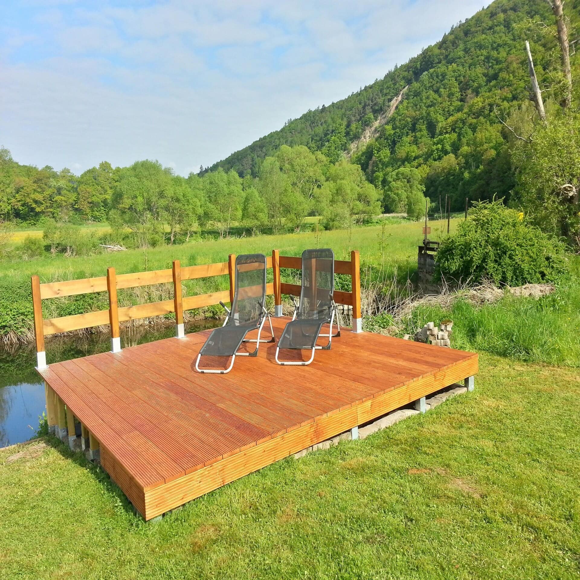 Holzdeck mit 2 Liegestühlen an einem kleinen Wasserlauf, dahinter die grüne Natur. Die Sonne scheint.