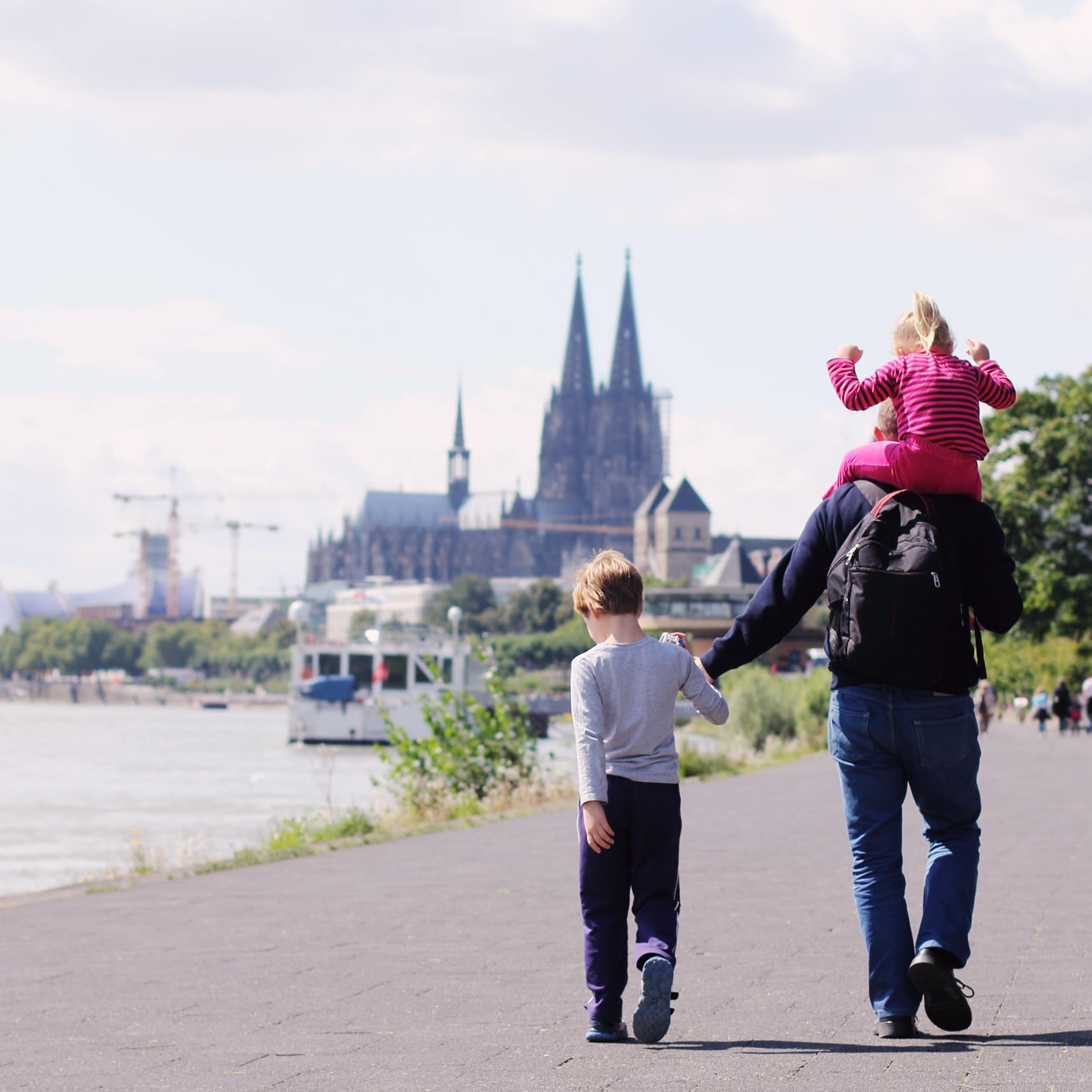 Spaziergang am Rheinufer: Vater mit Rucksack und kleinem Mädchen auf den Schultern und einem Jungen an der Hand.