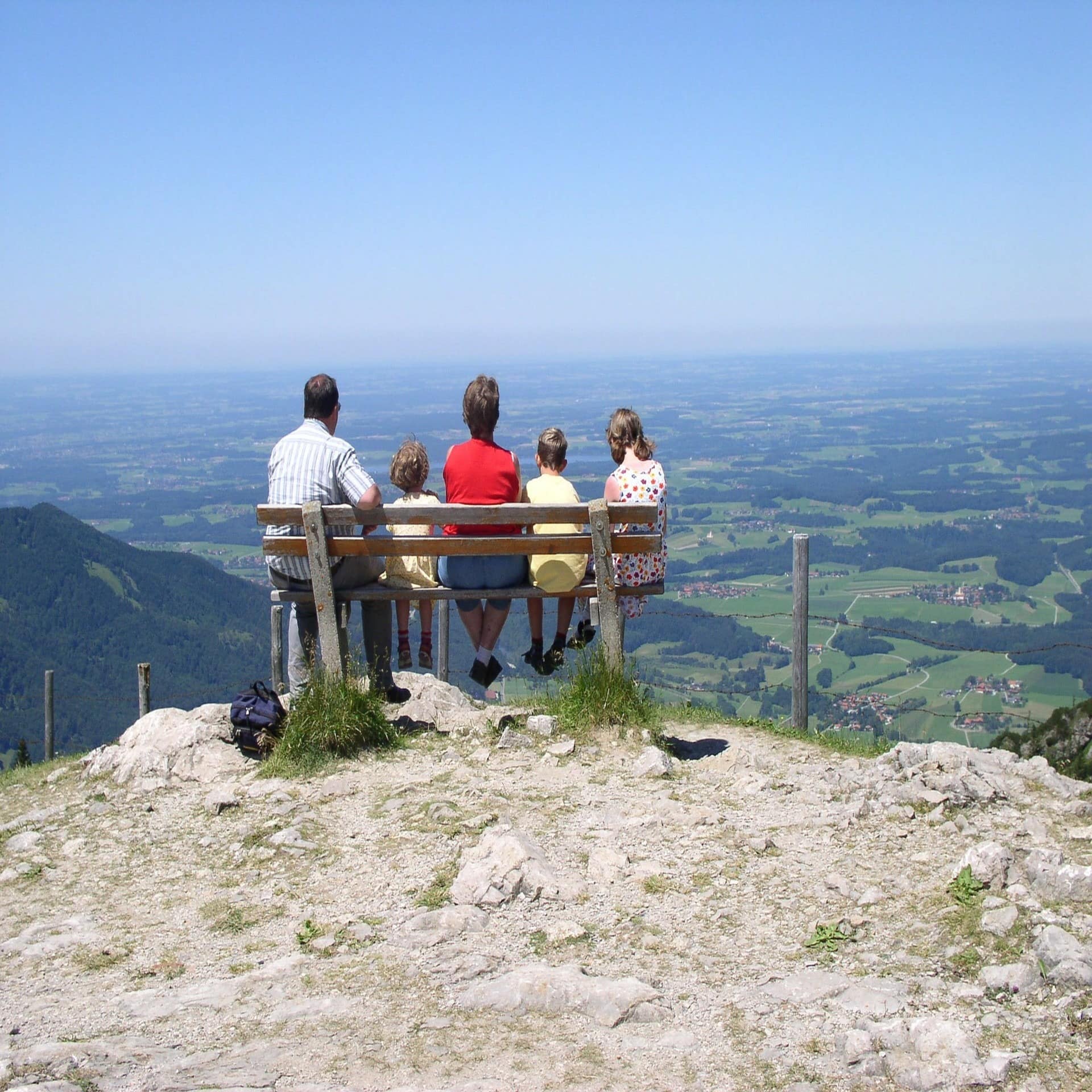 Rückenansicht: Eine Familie mit 3 Kindern sitzt auf einer Bergbank und blickt ins Tal.