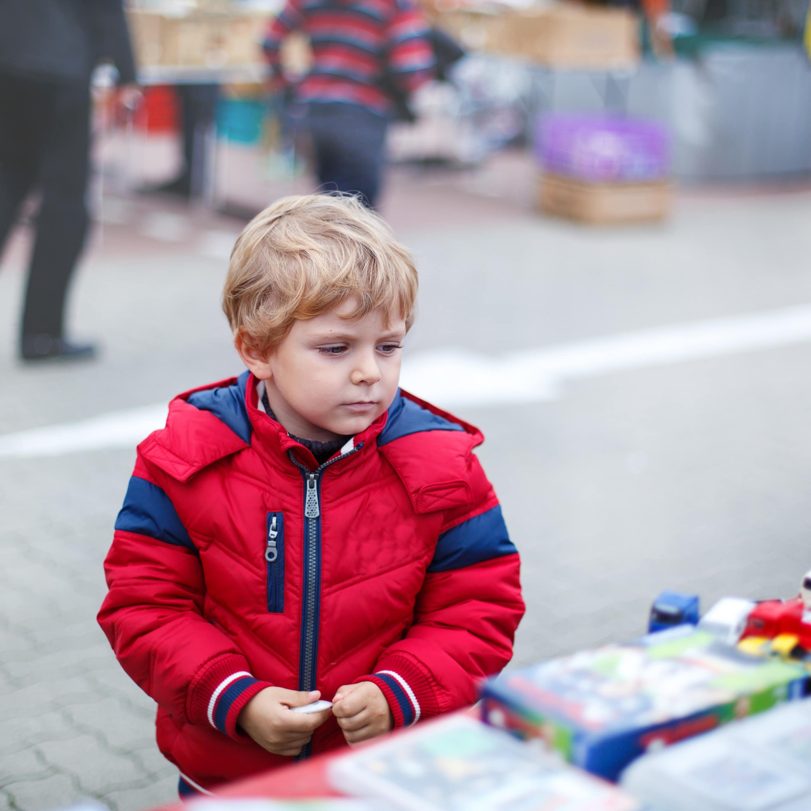 Kleiner blonder Junge mit roter Jacke betrachtet Spielzeug an einem Flohmarktstand.