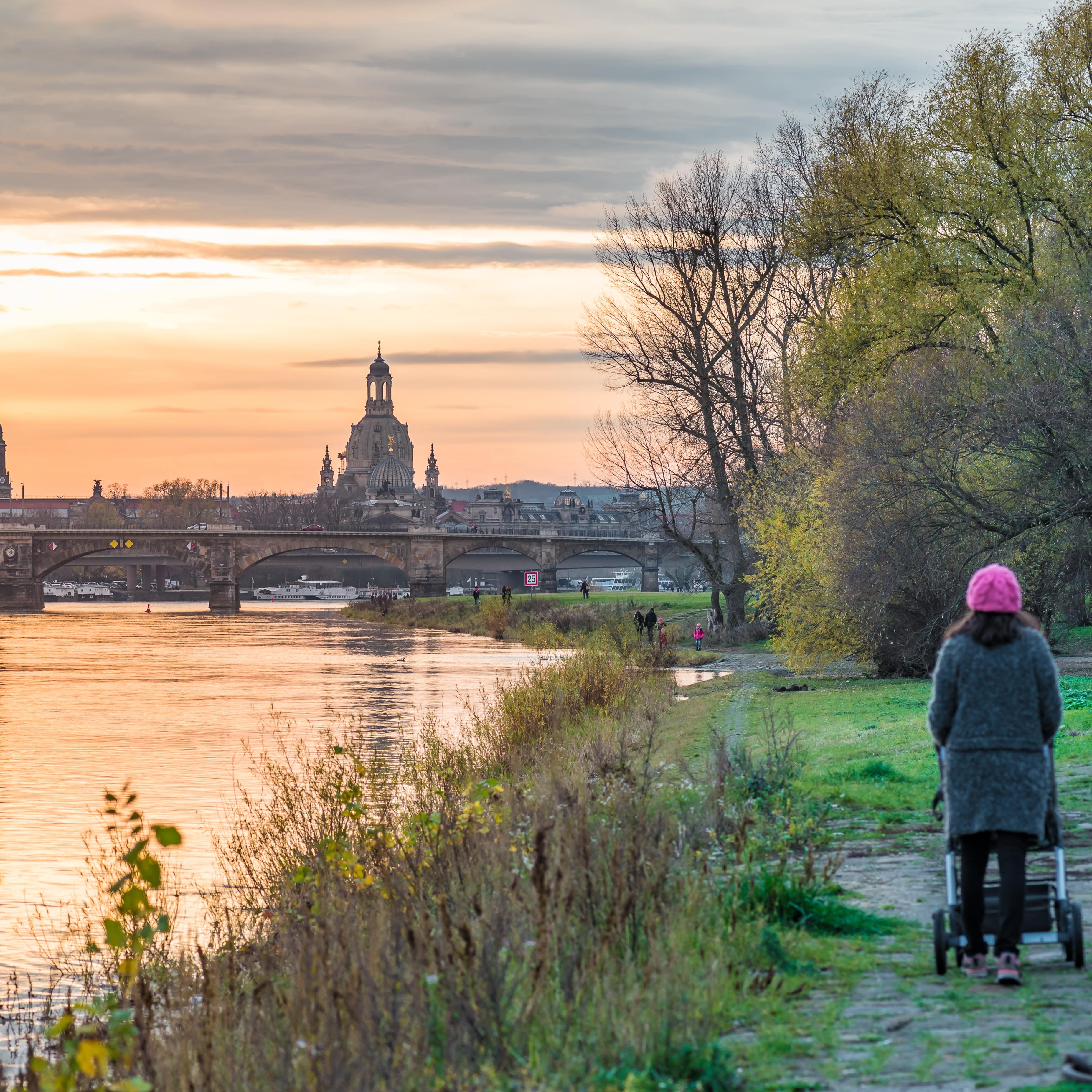Eine Frau mit Kinderwagen am Ufer der Elbe vor der Kulisse Dresdens