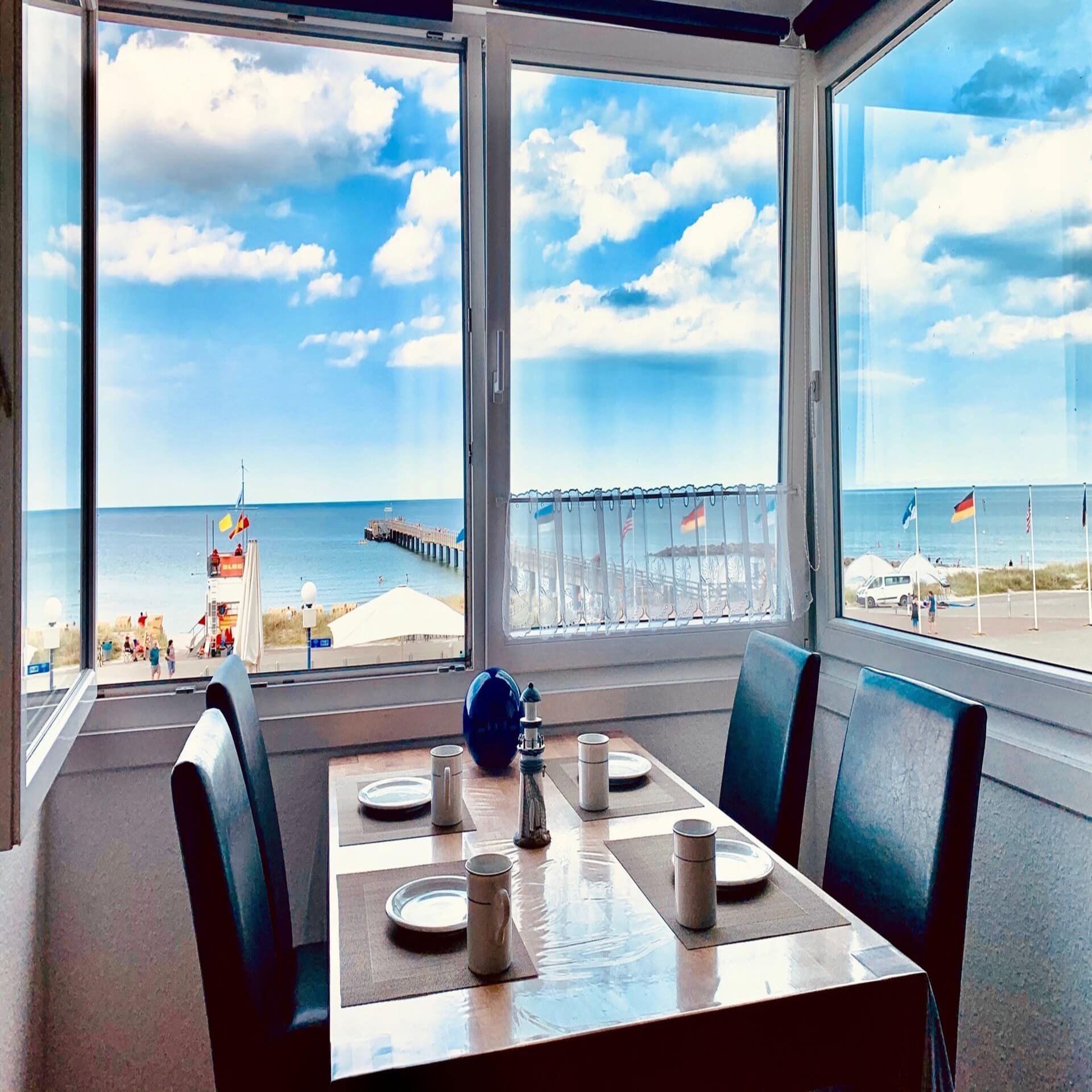 Esstisch für 4 Personen in einer Wohnung. Durch die Fenster sind das Meer und die Promenade zu sehen. 