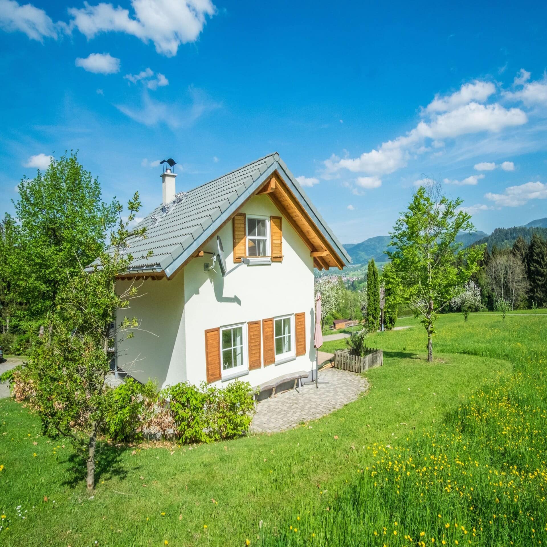 Weißes modernes Haus mit Garten im Grünen. Im Hintergrund einige Häuser. Die Sonne scheint. 