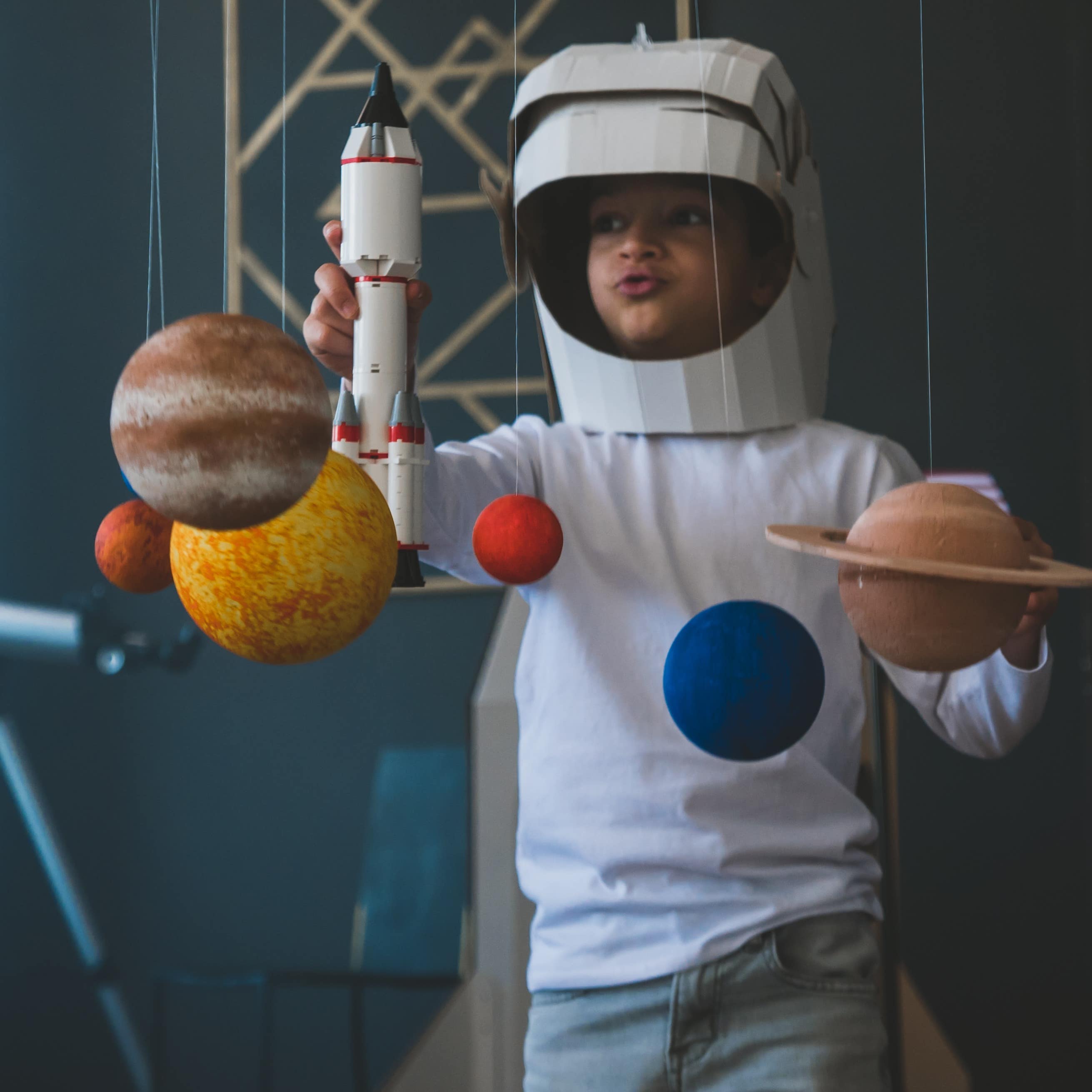Ein Junge mit Karton-Astronautenhelm spielt mit einer Spielzeugrakete und an Fäden hängenden Planeten.