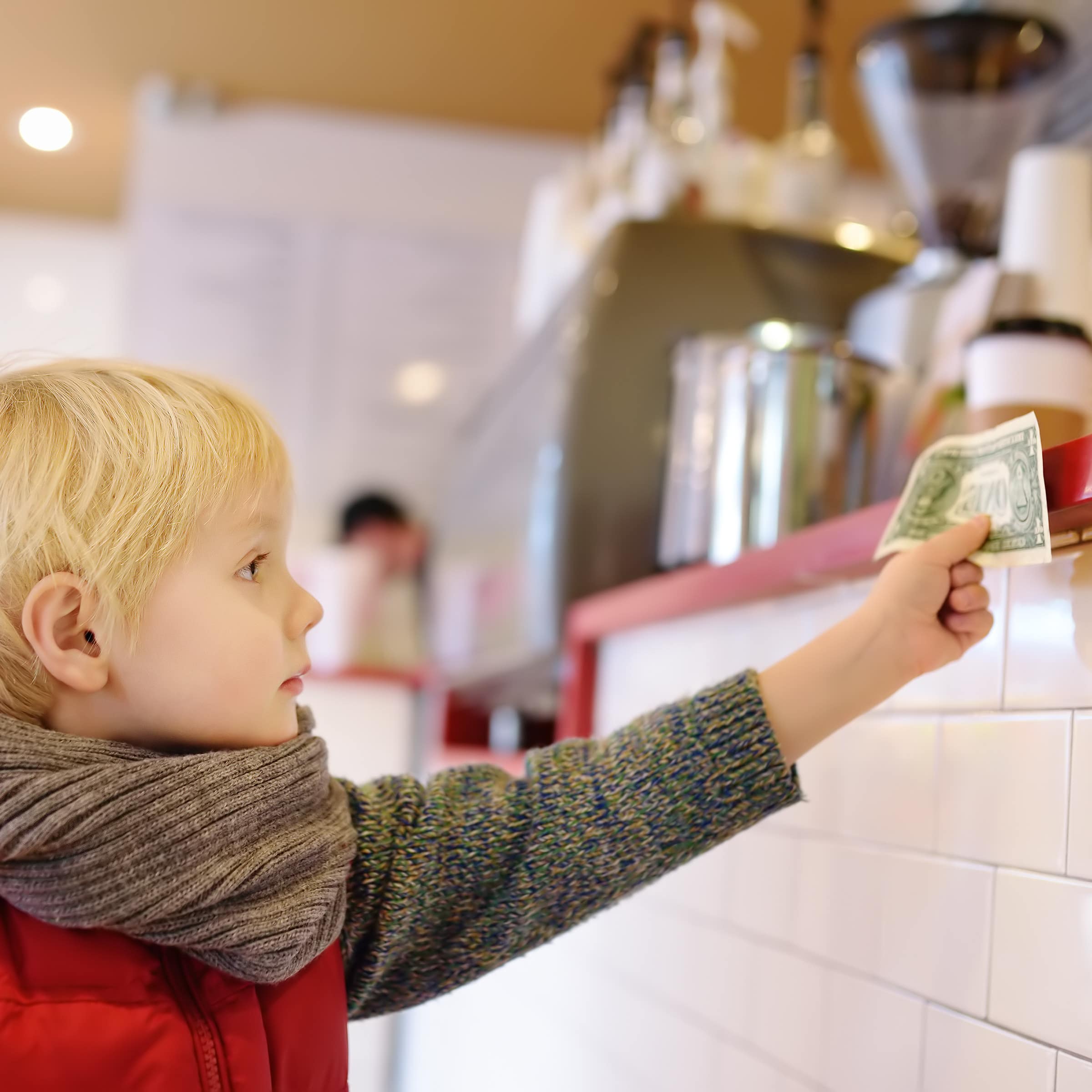 Ein kleiner blonder Junge hält einen Geldschein an den Tresen in einem Café. 