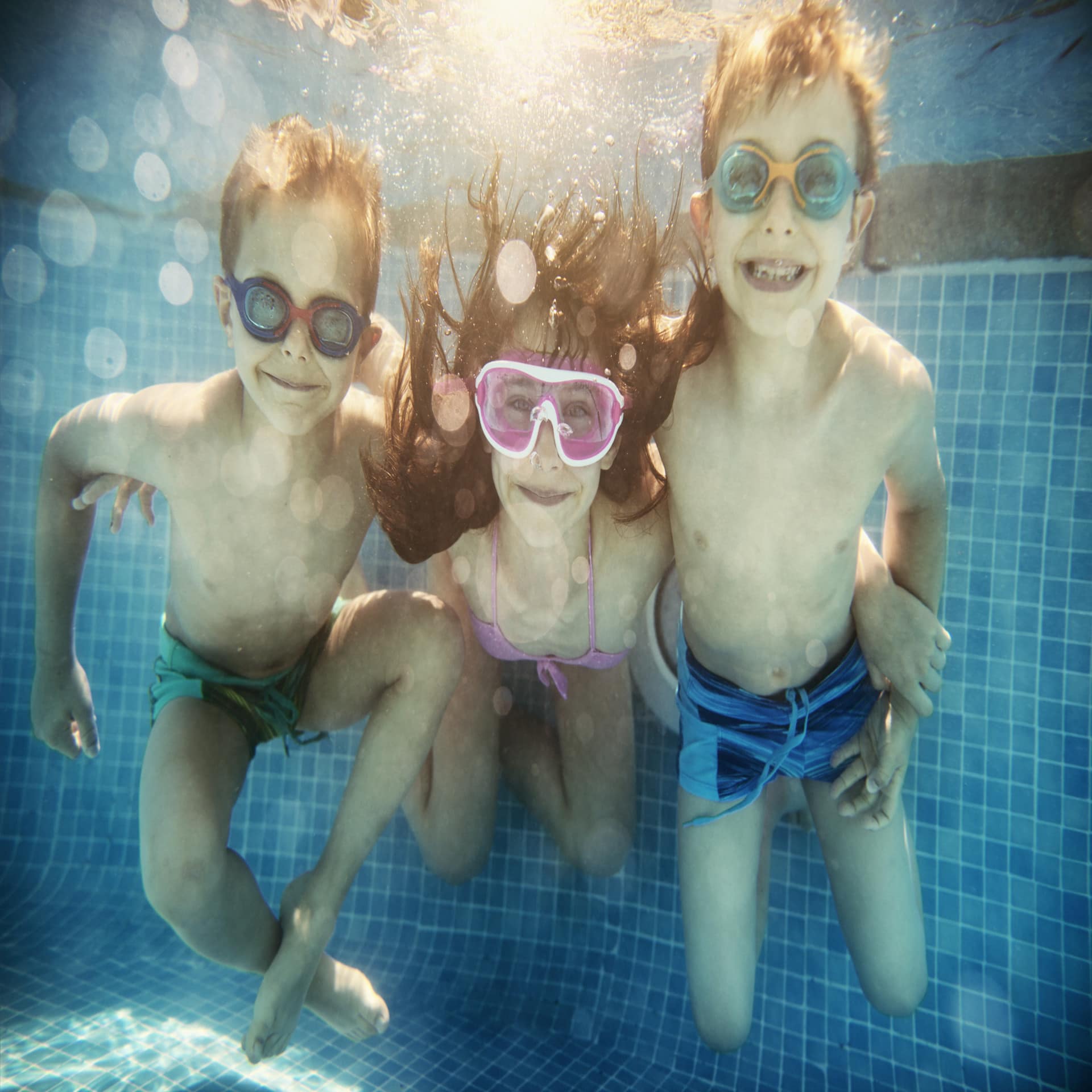 2 Jungs und 1 Mädchen in der Mitte tauchen unter Wasser, alle haben Schwimmbrillen auf.