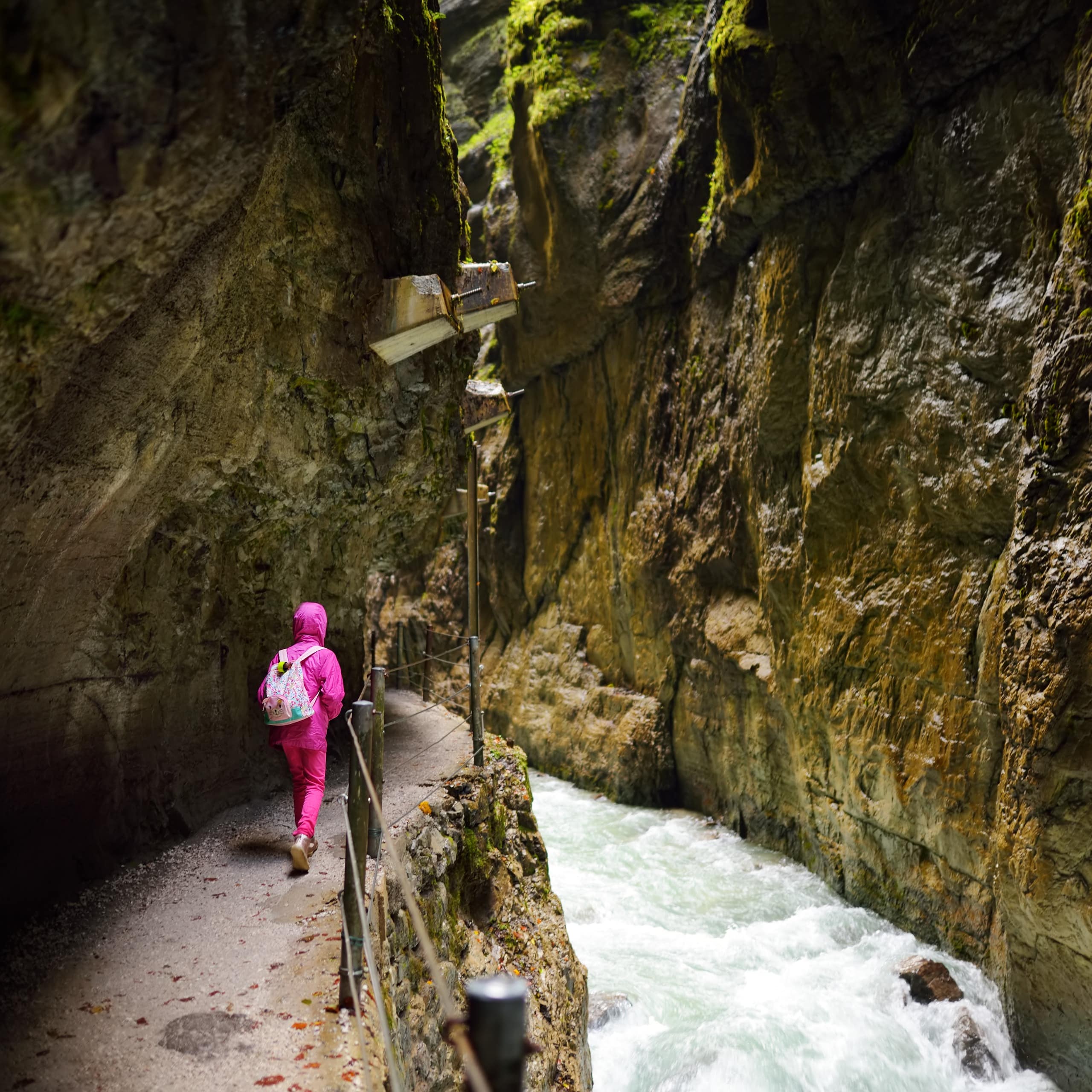 Rückenansicht: Ein Mädchen in pinker Kleidung mit rosa Rucksack läuft durch die Partnachklamm. Rechts Wasser.