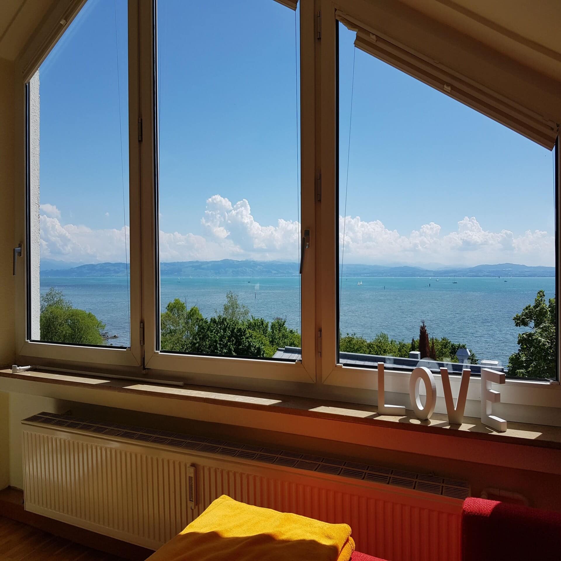 Blick aus einem Fenster über ein paar Bäume auf den Bodensee. Die Sonne scheint.