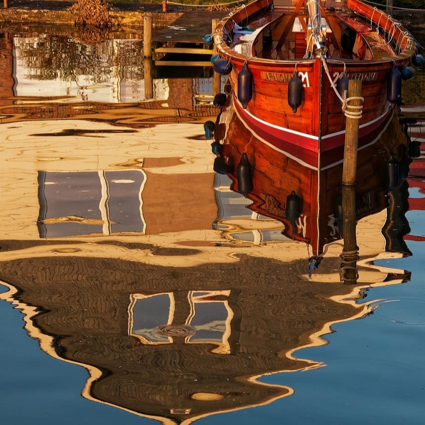 Ein Ruderboot an einem Steg, im Wasser spiegelt sich ein Haus. 