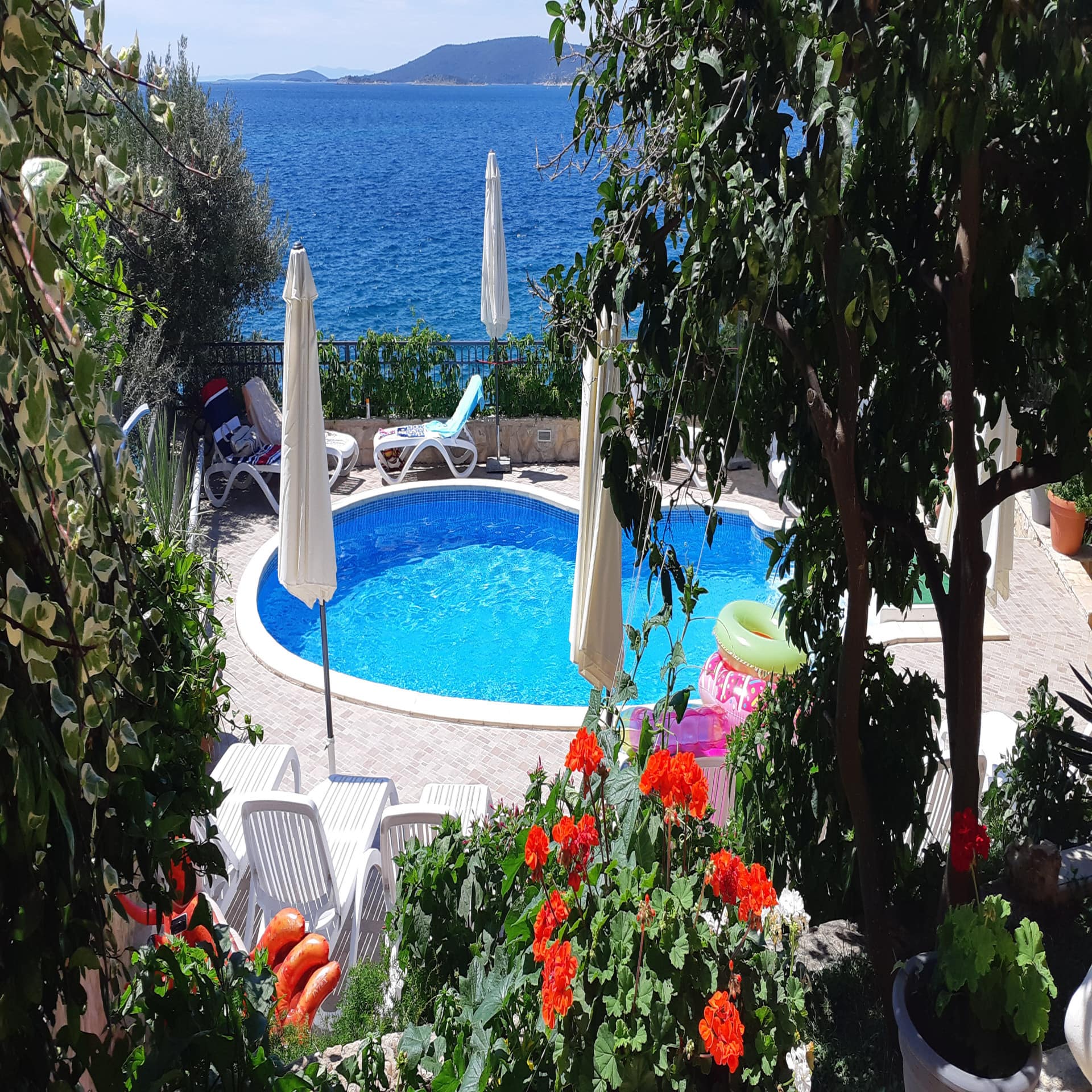 Versteckter Garten mit Pool und Liegestühlen sowie Blick auf das Meer. Die Sonne scheint.