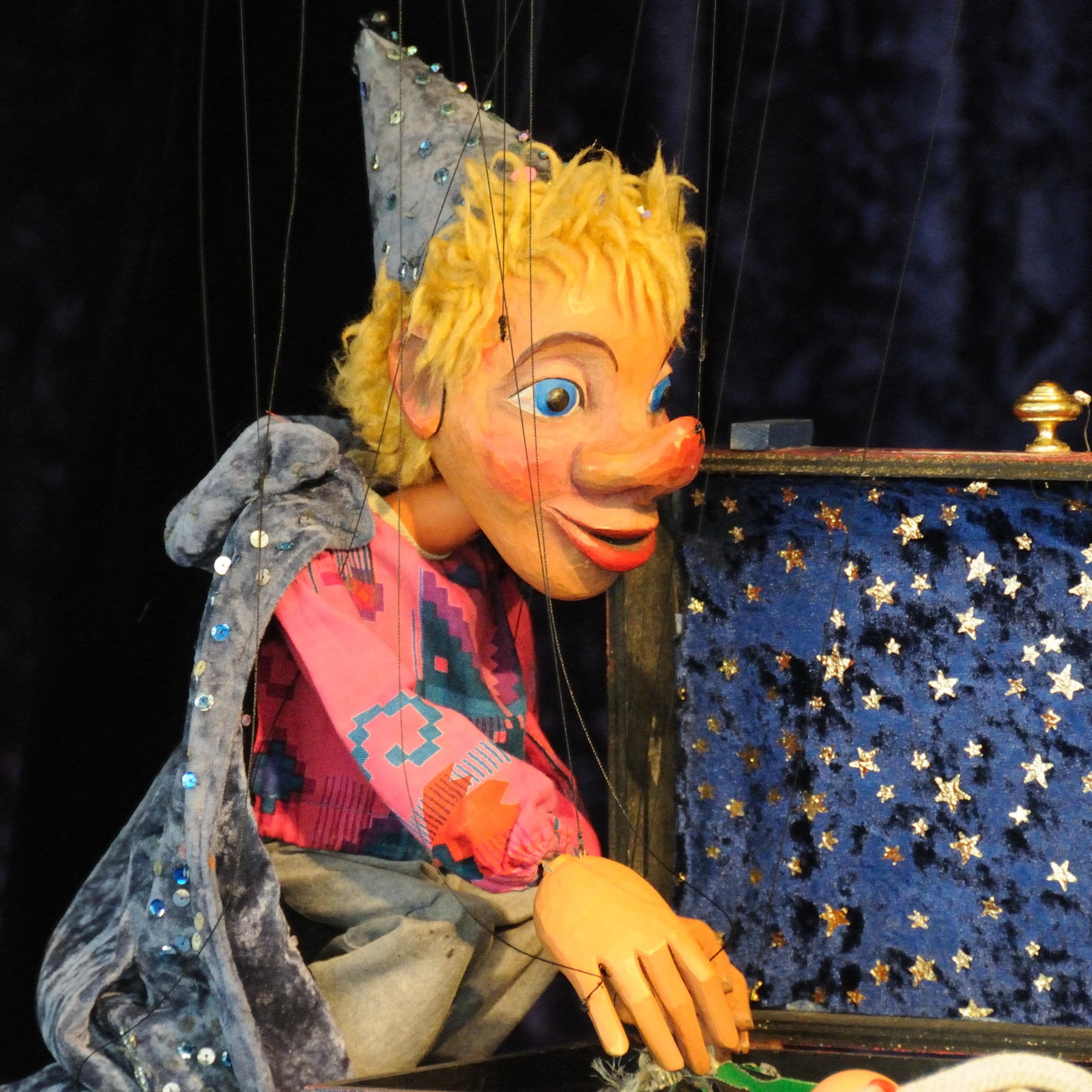 Eine Kasperle-Marionette mit blauem Umfang an einem geöffneten Koffer mit blauem Sternenbezug.