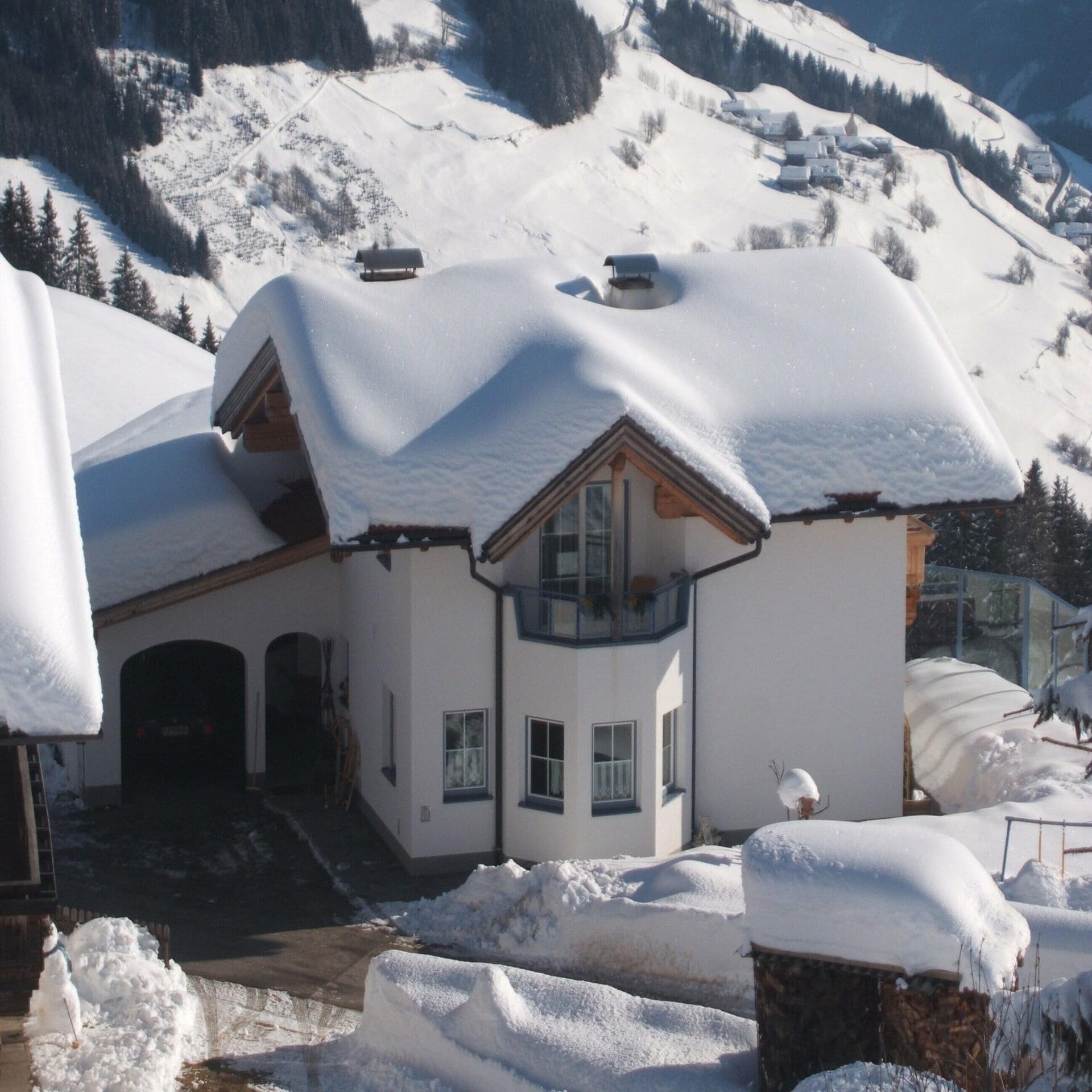 Ein weißes Haus in der verschneiten Landschaft in Tirol. Im Hintergrund einige Häuser. Die Sonne scheint. 