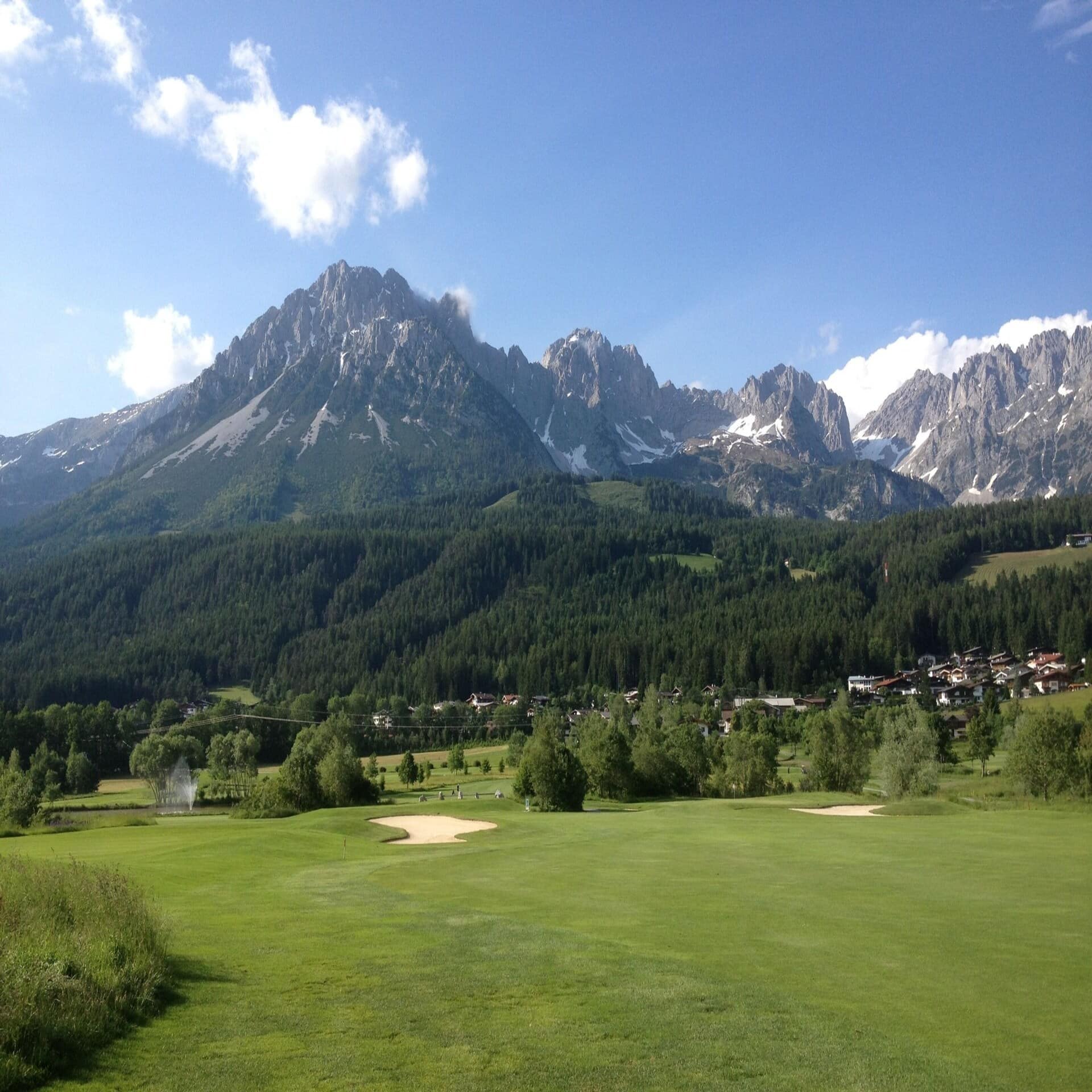 Blick über einen Golfplatz und ein Dorf unterhalb des Wilden Kaisers im Sommer.