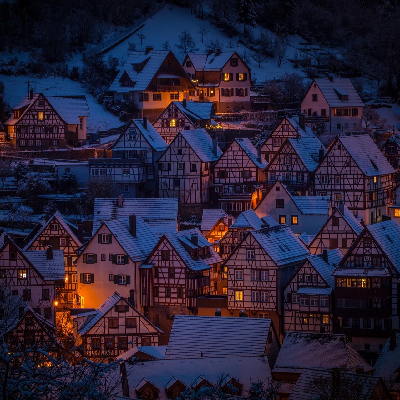 Ein verschneites Dorf am Abend am Fuße eines bewaldeten Bergs, einige der Fachwerkhäuser sind erleuchtet. 