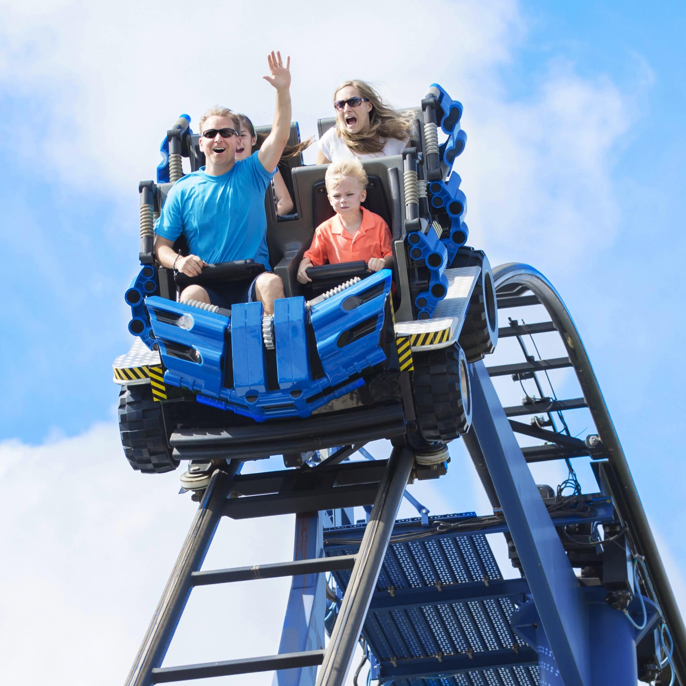 Vater, Mutter und 2 Kinder sitzen in einer Achterbahn, die steil bergab fährt. Vater reckt die Hand hoch.