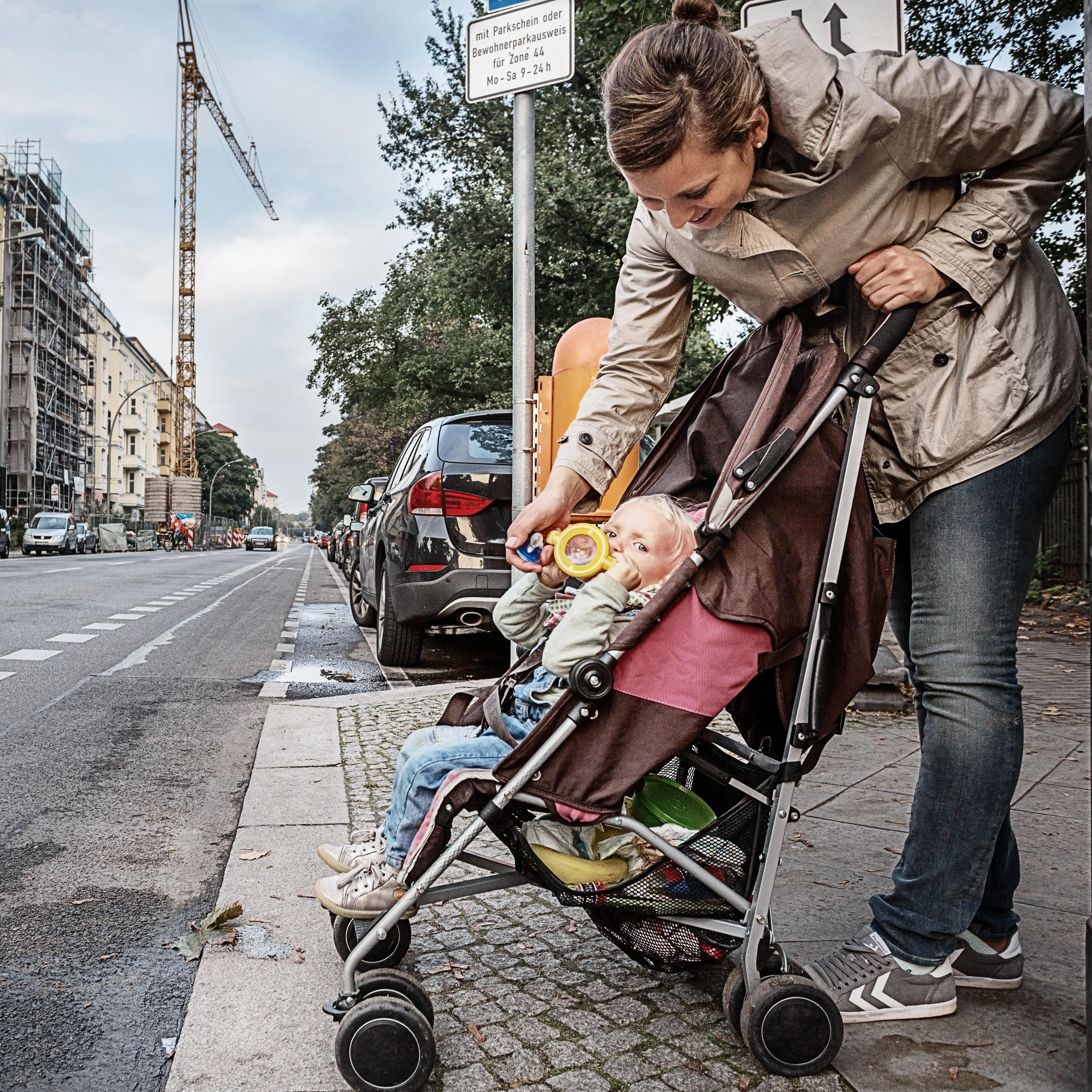 Mutter und Kleinkind im Buggy warten am Straßenrand, um die Straße zu überqueren. Die Mutter gibt dem Kind einen Becher.