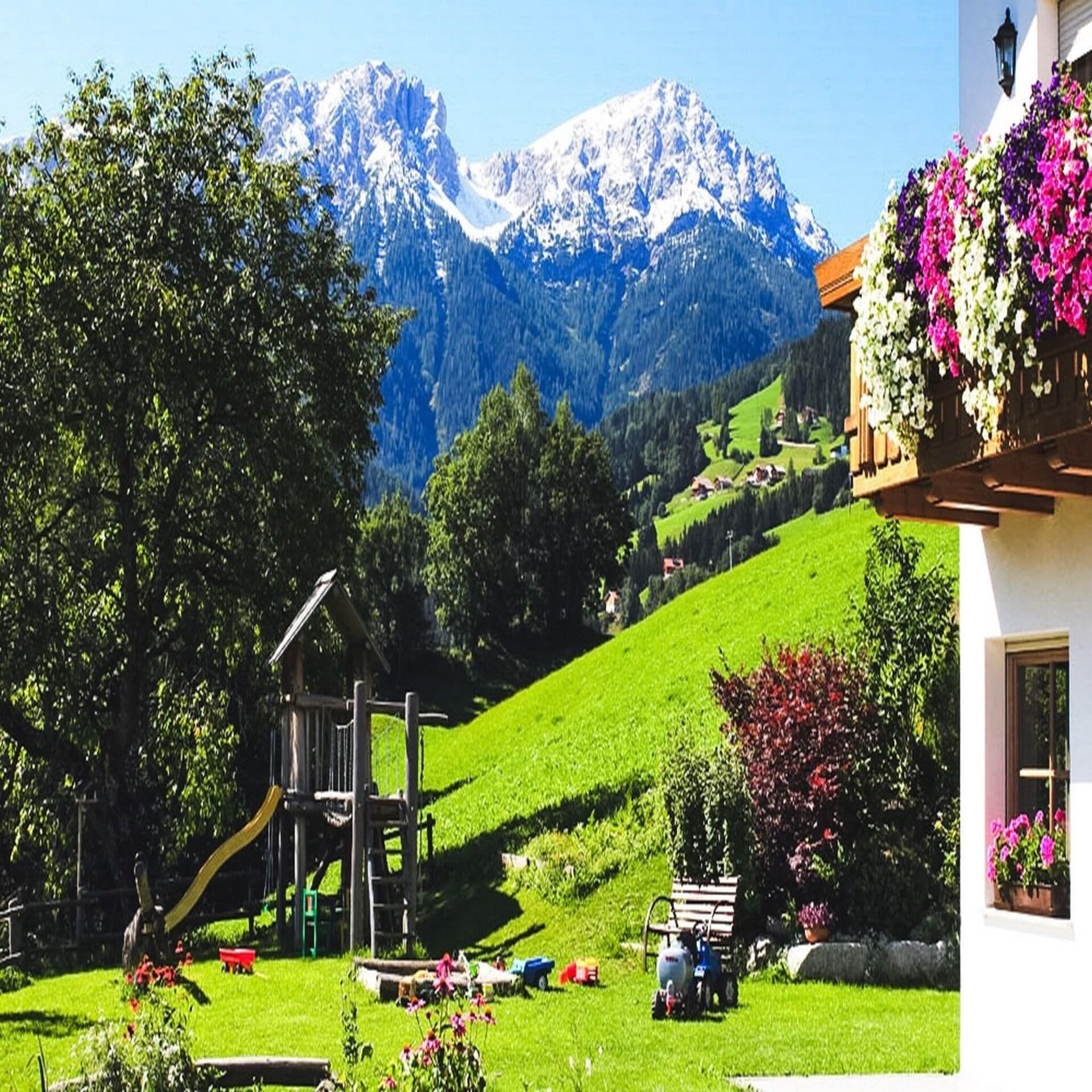 Ein Garten mit Spielplatz, rechts ist ein Teil eines Hauses zu sehen. Die Berge und Wiesen im Hintergrund. 