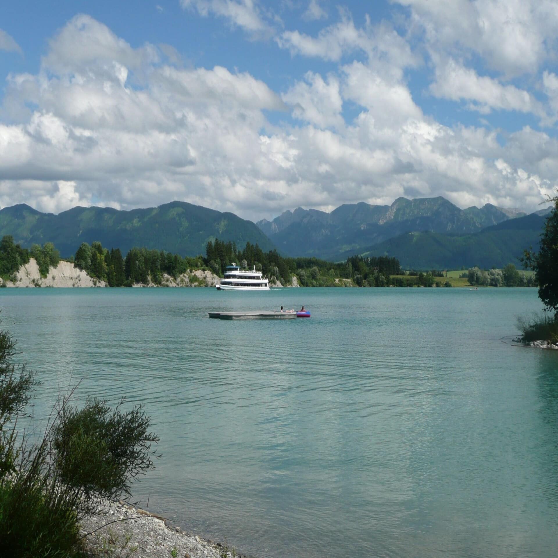 Ein Ausflugsschiff fährt auf den Forggensee, im Hintergrund Wiesen und bergige Wälder. 