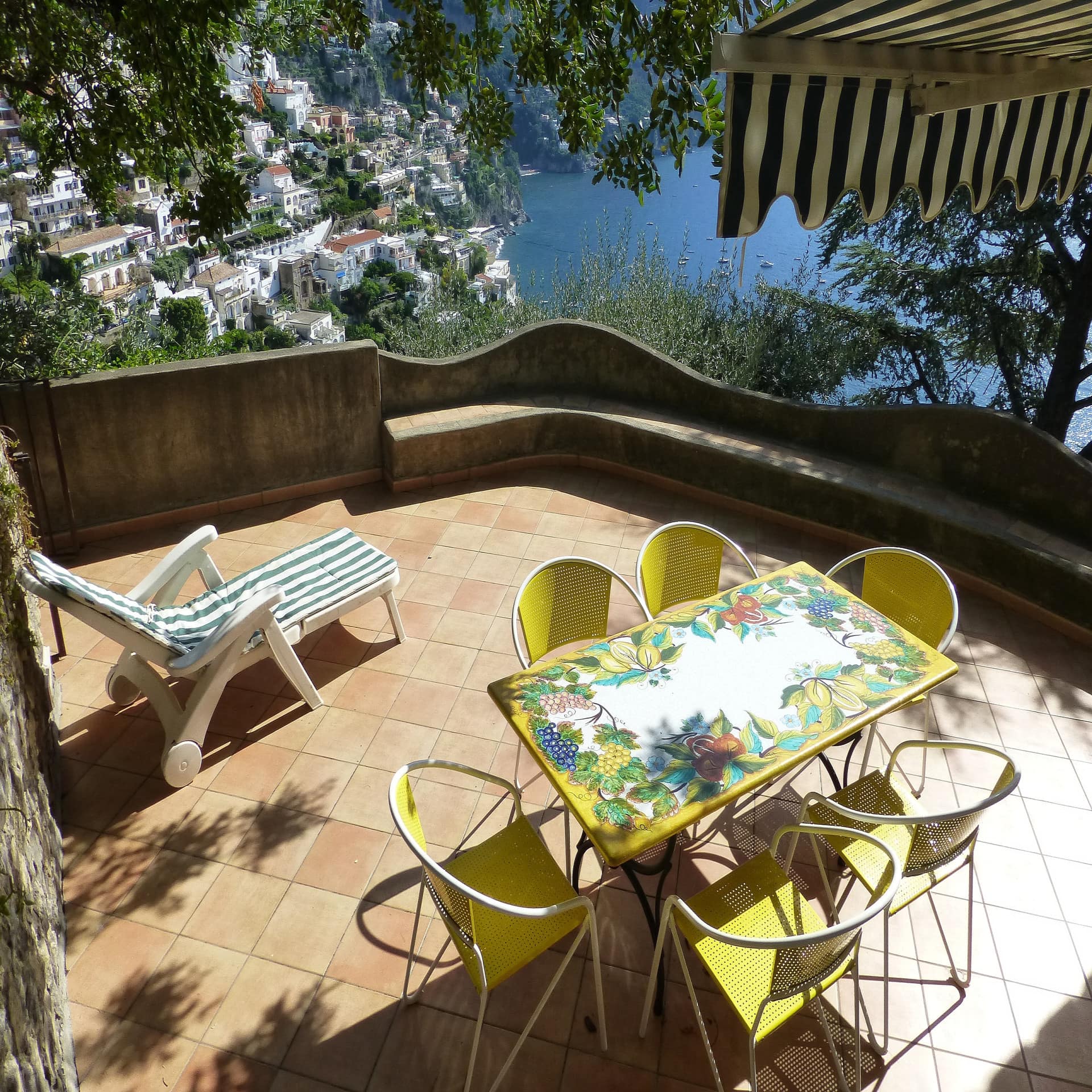 Gelber Tisch und Stühle und eine Sonnenliege auf einer Terrasse mit Blick über einen Ort an der Amalfiküste.  