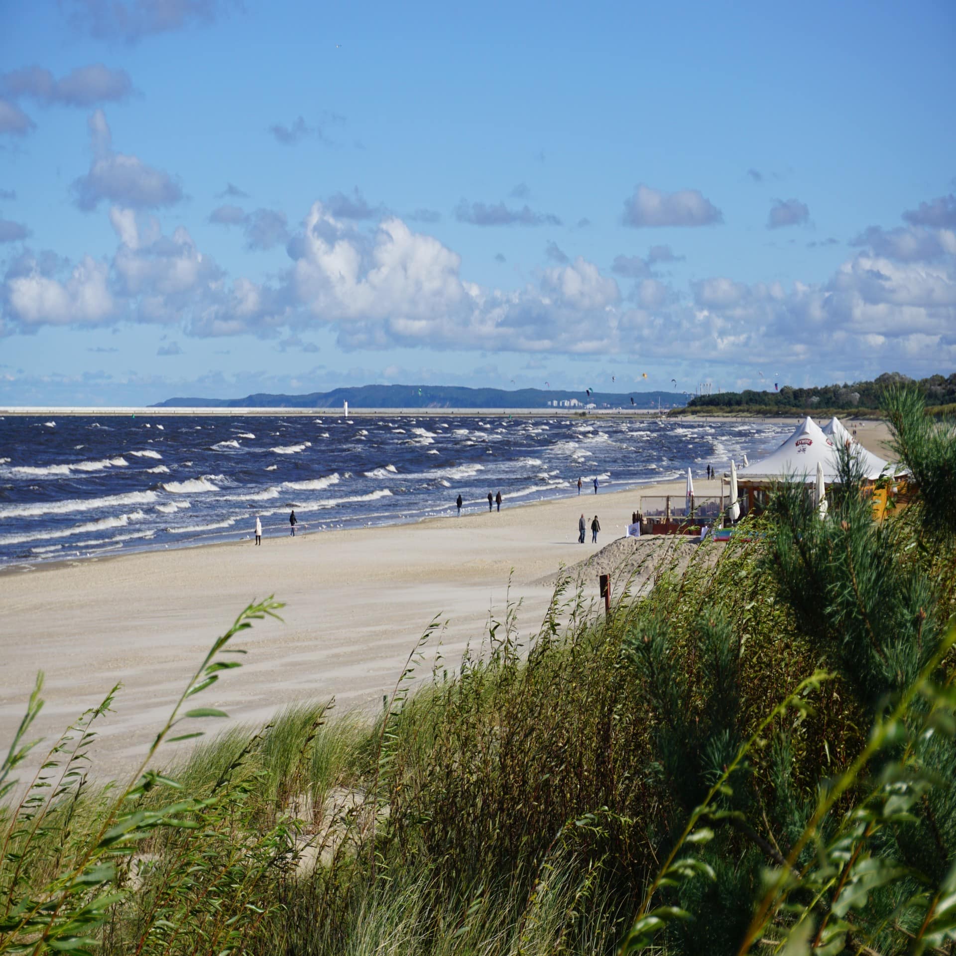 Blick über die Dünen auf den Strand und das raue Meer von Swinemünde. Leute gehen am Strand spazieren.